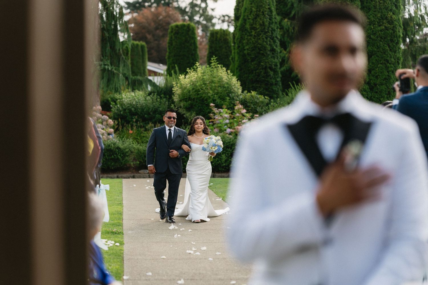 Bride walks down the aisle with escort as groom awaits at outdoor wedding ceremony.