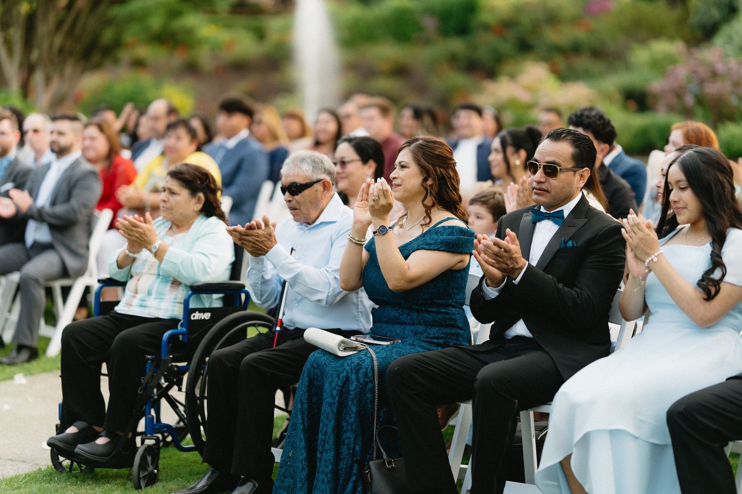 Wedding guests applaud during outdoor garden ceremony, including guest in wheelchair.