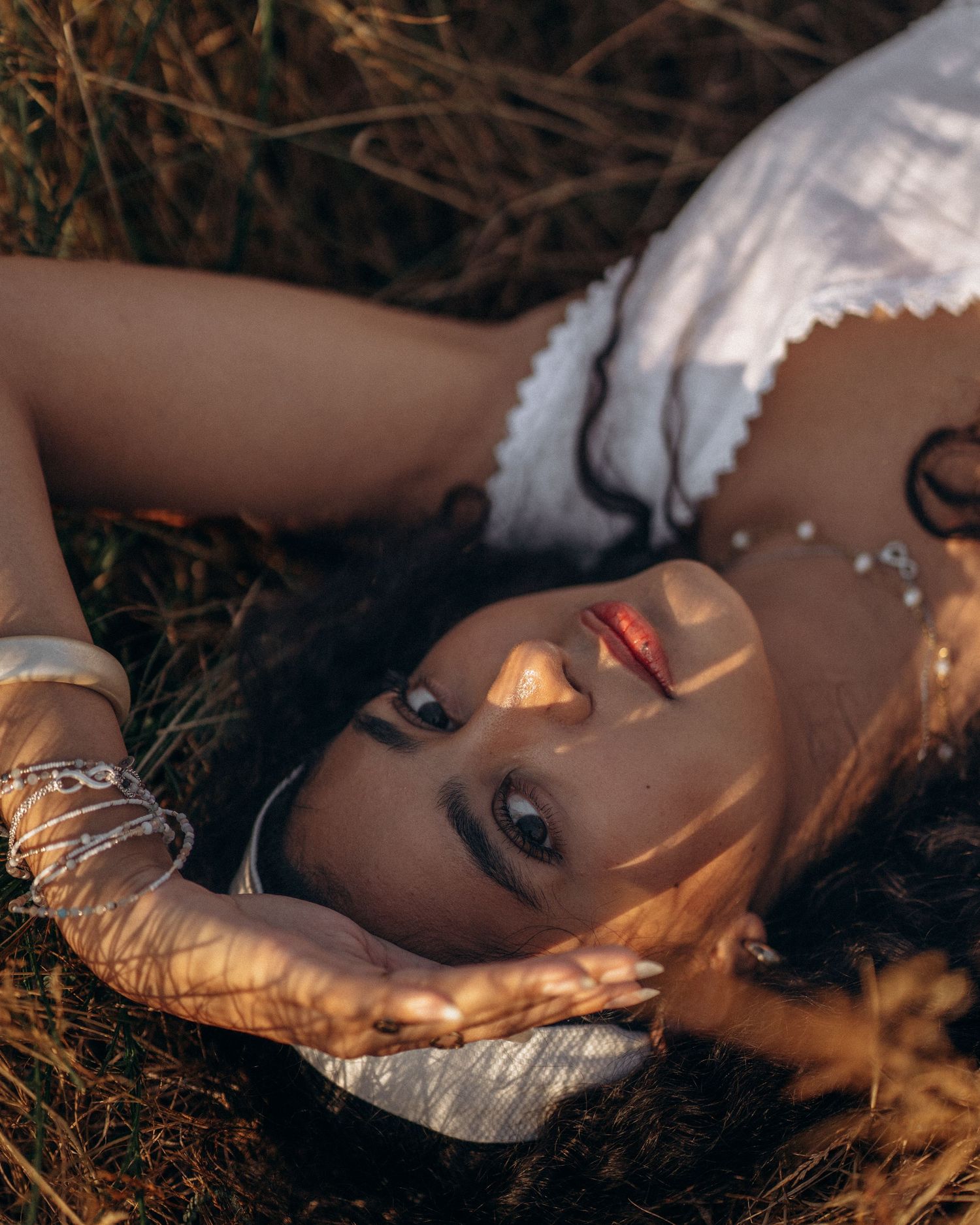 Woman in white dress lying in dry grass field, bathed in warm golden sunlight with silver jewelry.