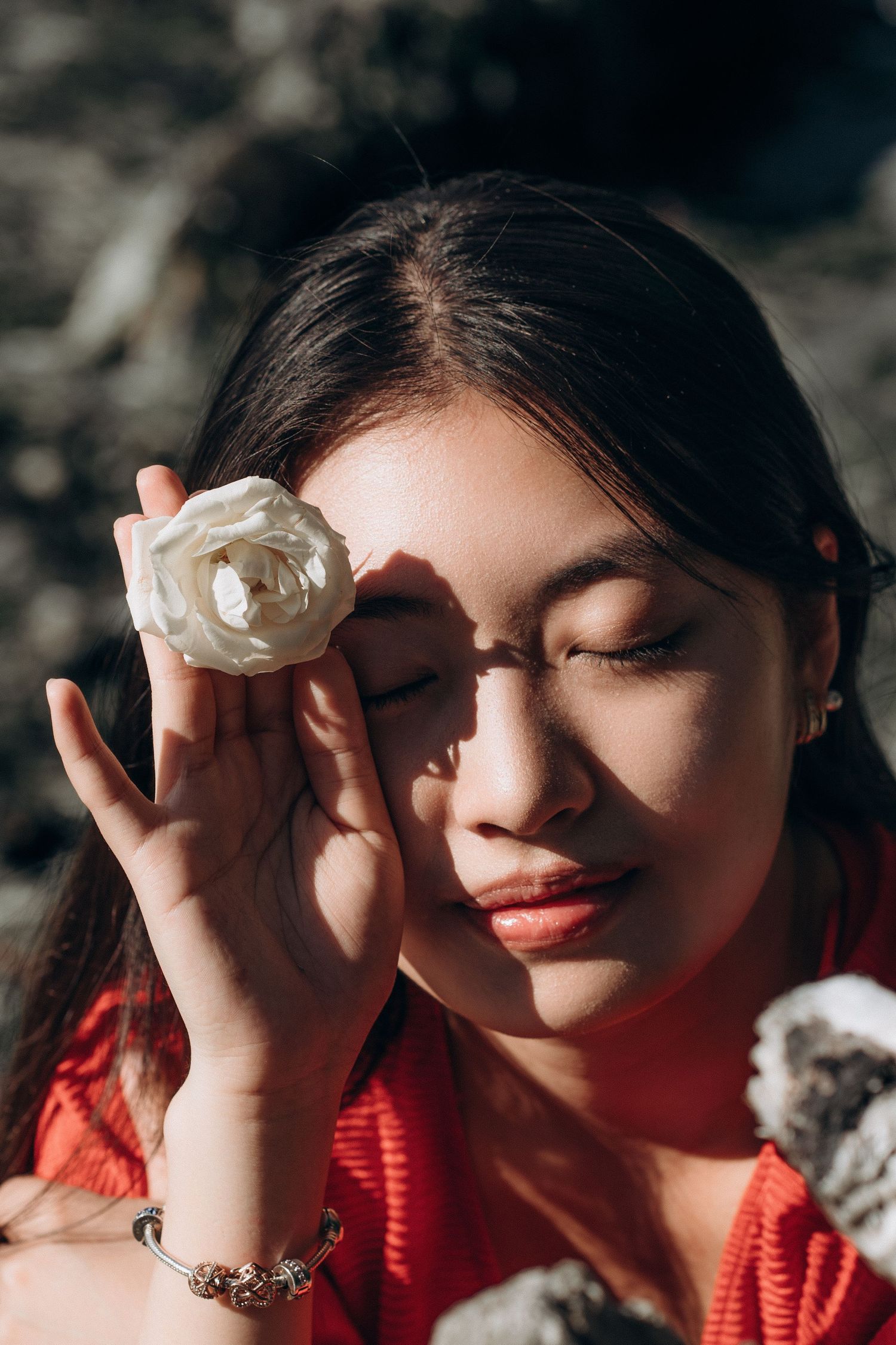 Close-up of young woman in red holding a white rose over her eye in sunlight.