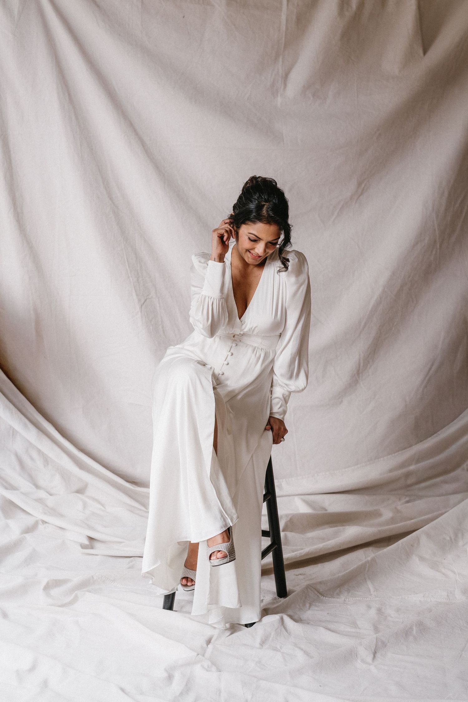Bride in elegant white satin jumpsuit sits on stool against draped linen fabric backdrop.