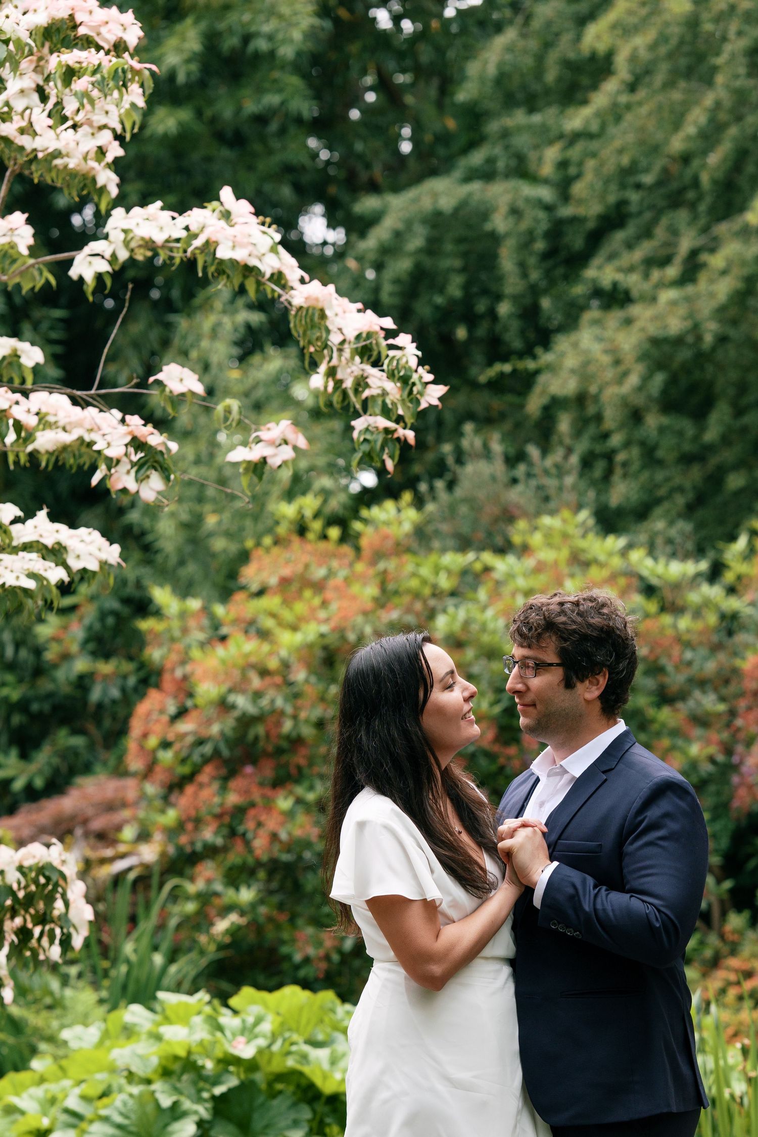 Couple gazing at each other under blooming white flowers in a garden, woman in white dress holding man's lapel.