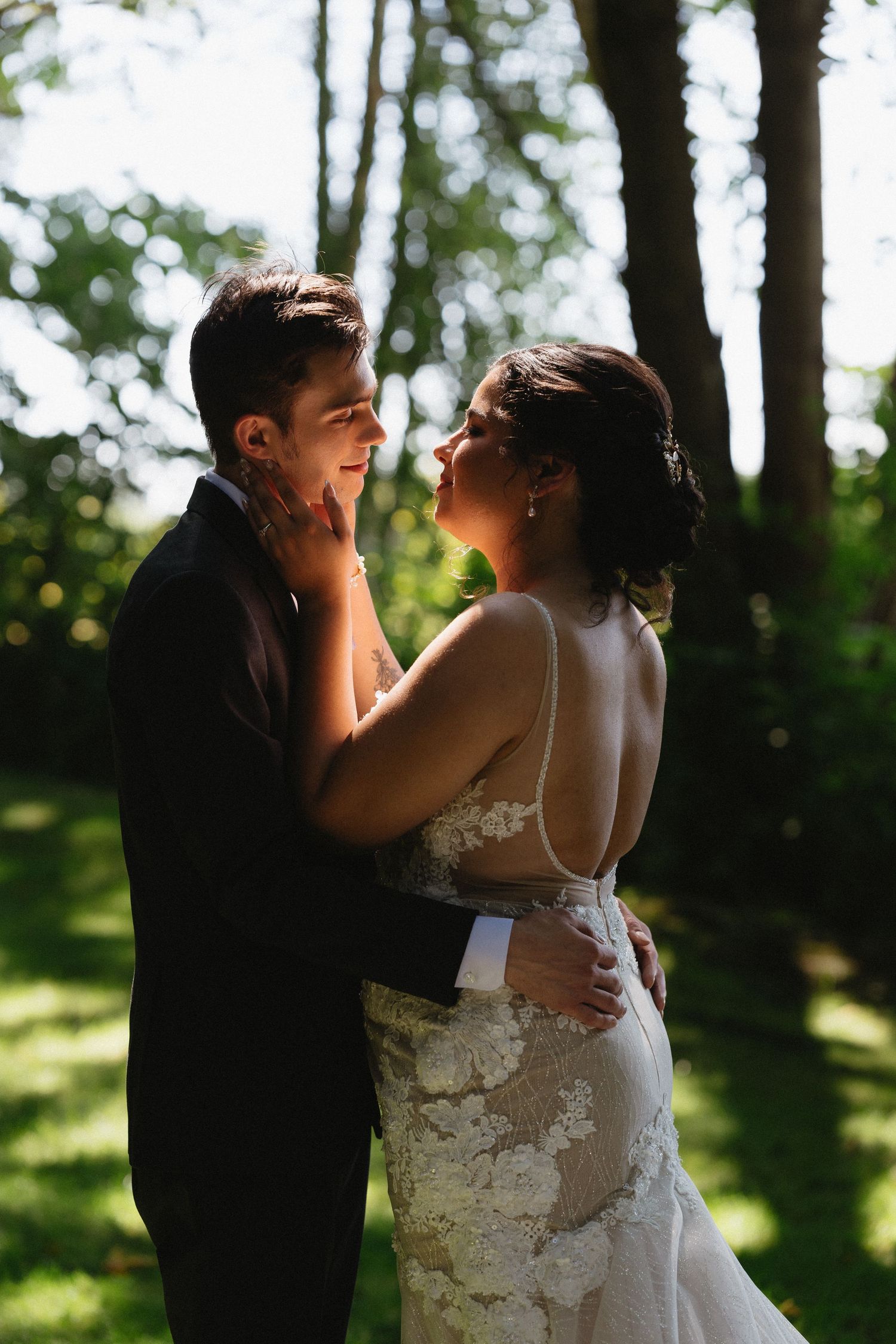 Bride in backless lace gown tenderly holds groom's face in a sunlit outdoor garden setting.
