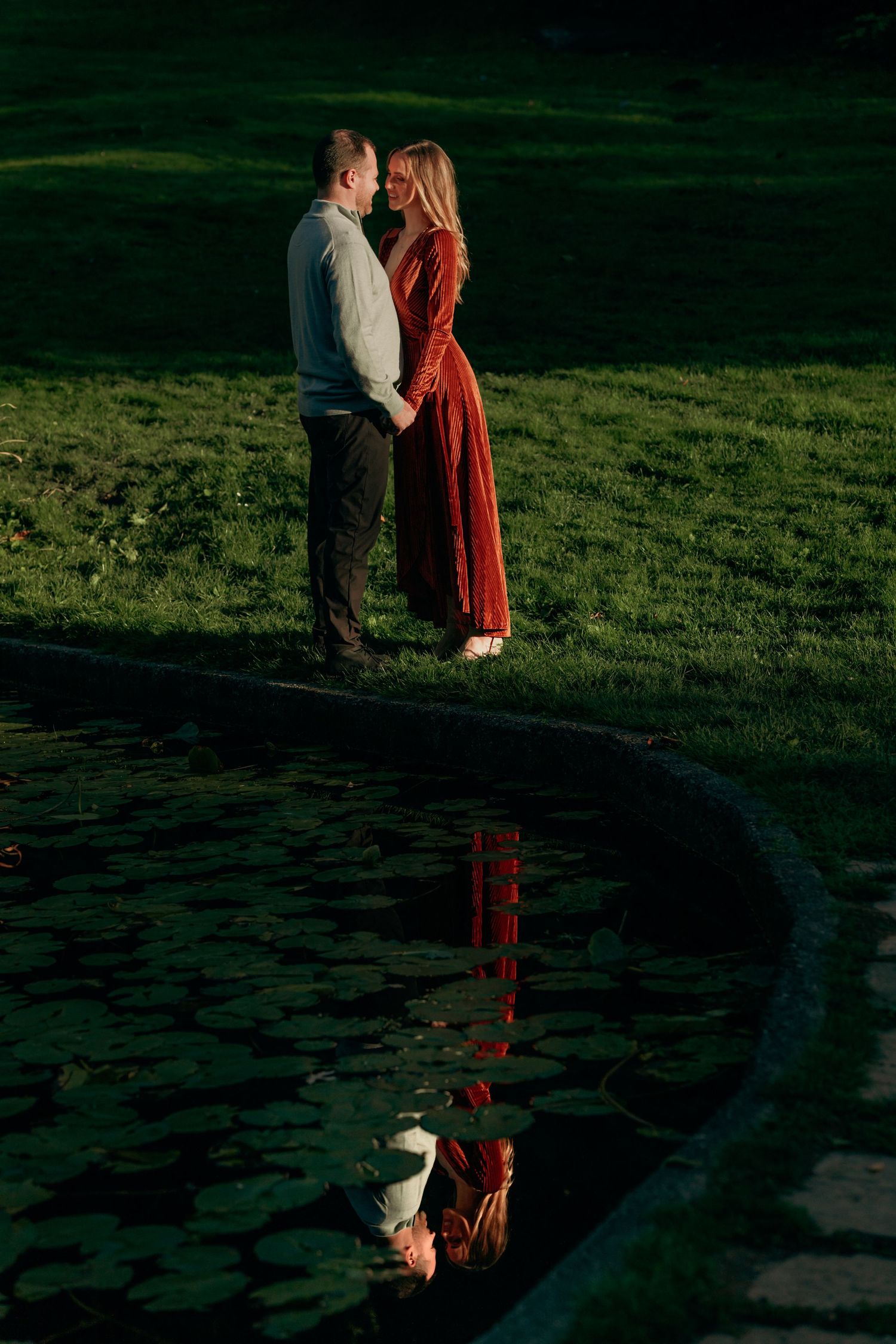 Couple holding hands by a lily pond at sunset, with their reflection visible in the water.