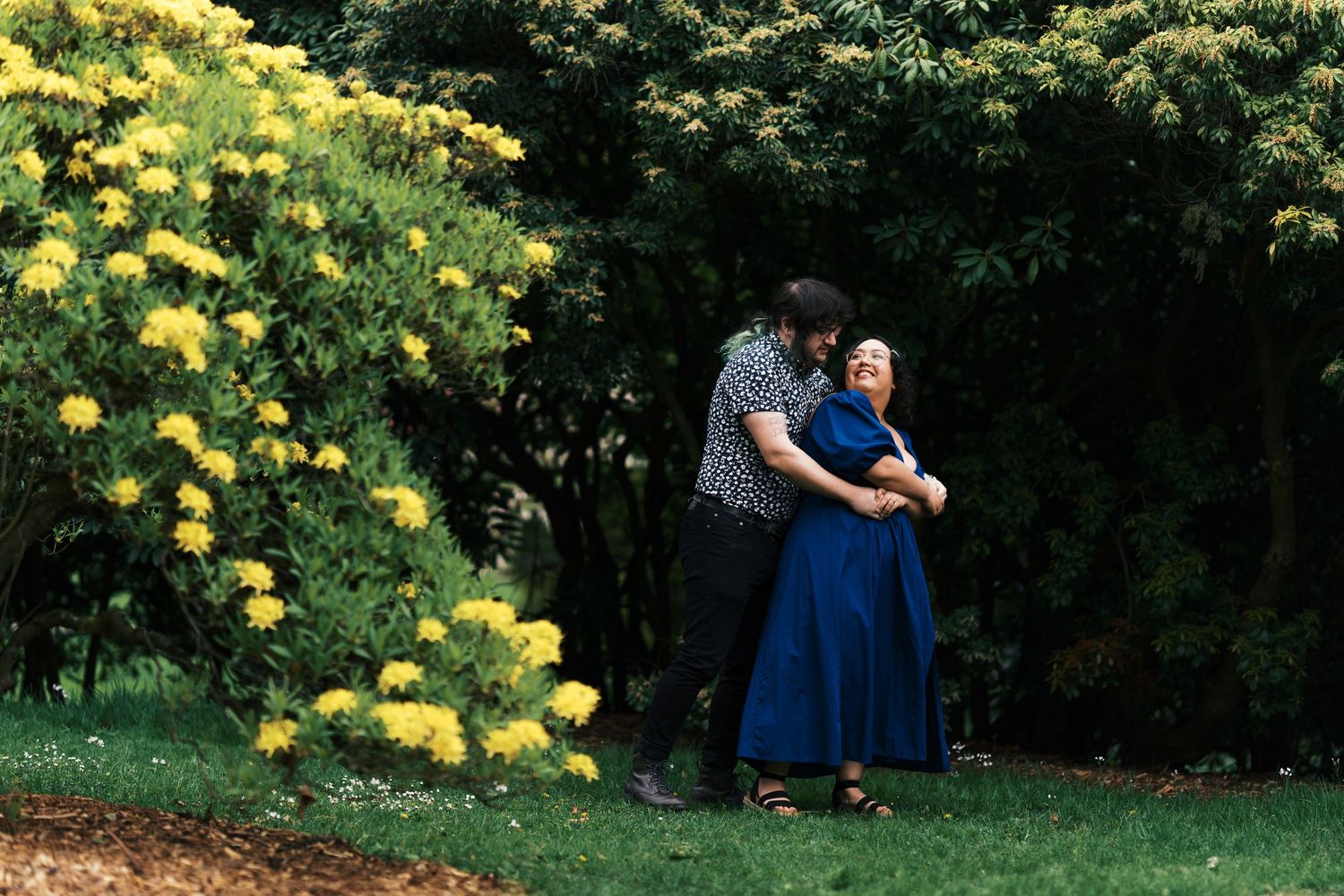 Couple sharing a tender embrace near vibrant yellow flowering bushes in a lush green garden.