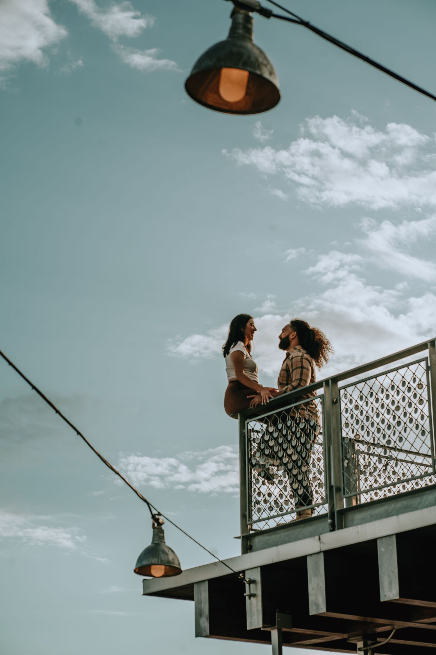 Romantic couple sharing an intimate moment on an elevated outdoor balcony against a blue sky with hanging vintage lights.