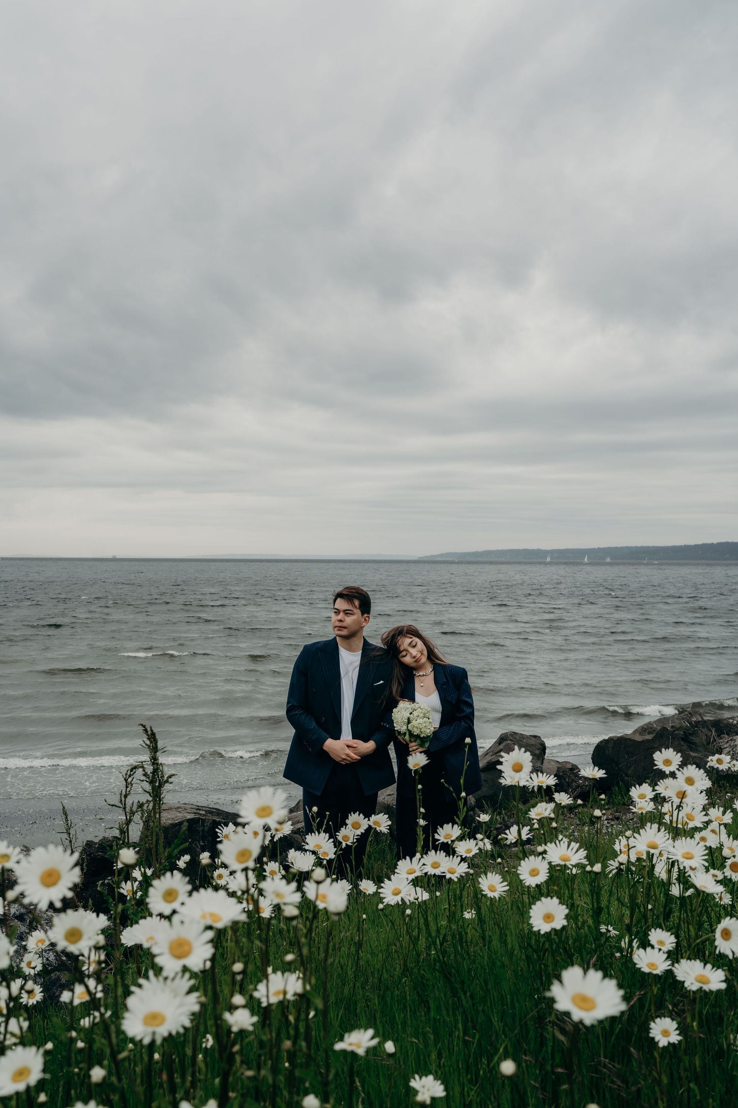 Couple in navy suits standing among white daisies with rocky ocean shoreline behind them.