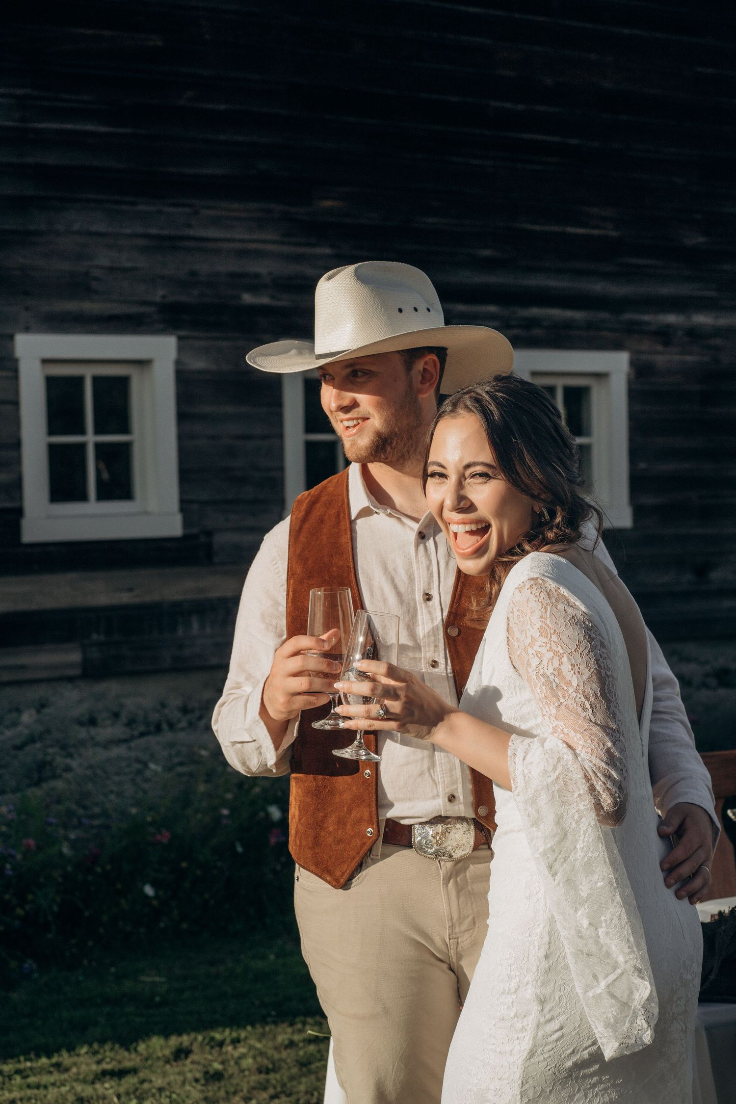 Happy bride and groom toasting with champagne flutes outside a rustic dark wood barn at golden hour.