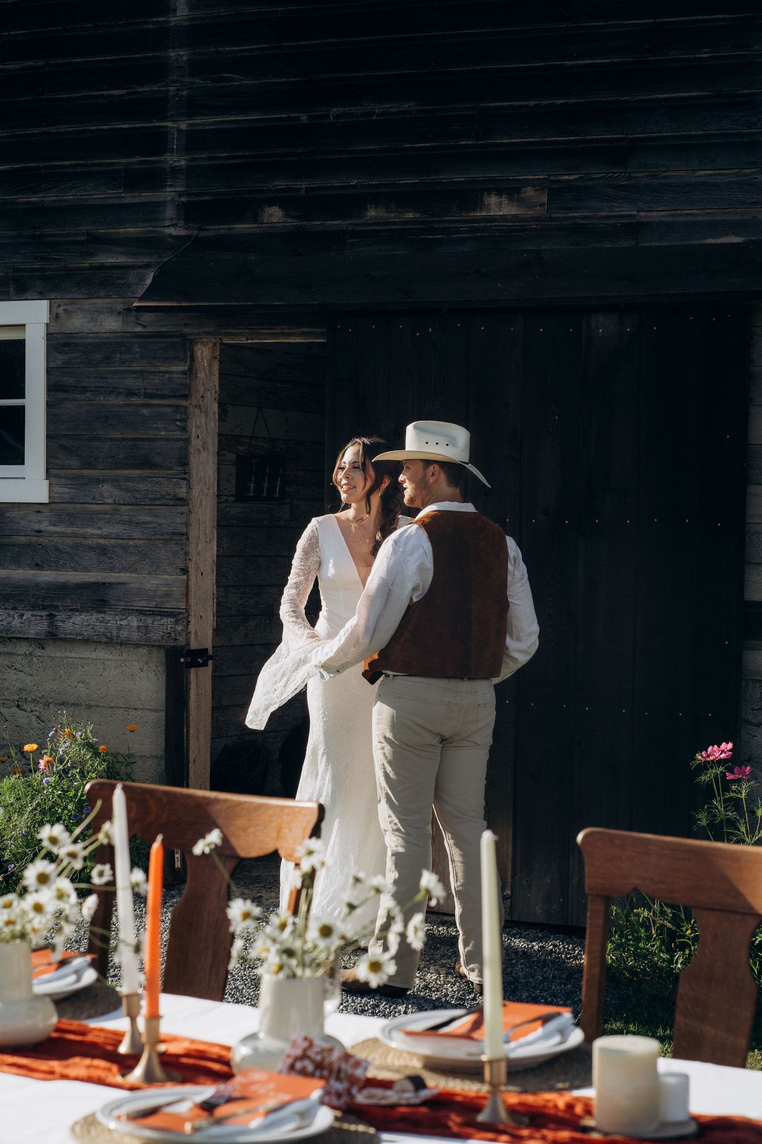 Couple sharing a romantic moment outside a weathered barn, groom in cowboy hat and brown vest.