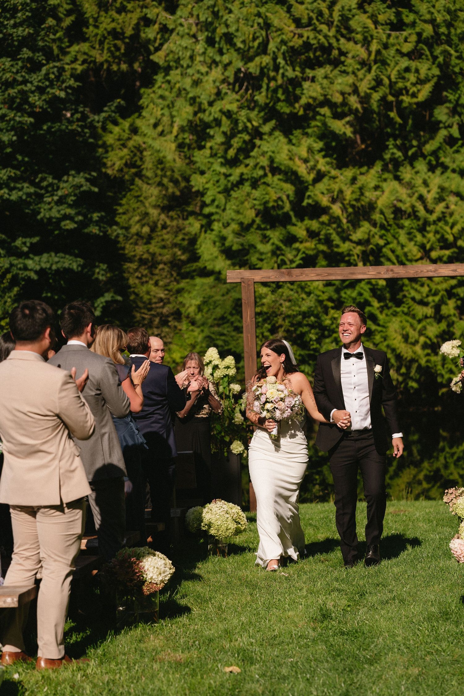 Newlyweds joyfully walking back down the aisle as guests applaud at outdoor wedding ceremony.