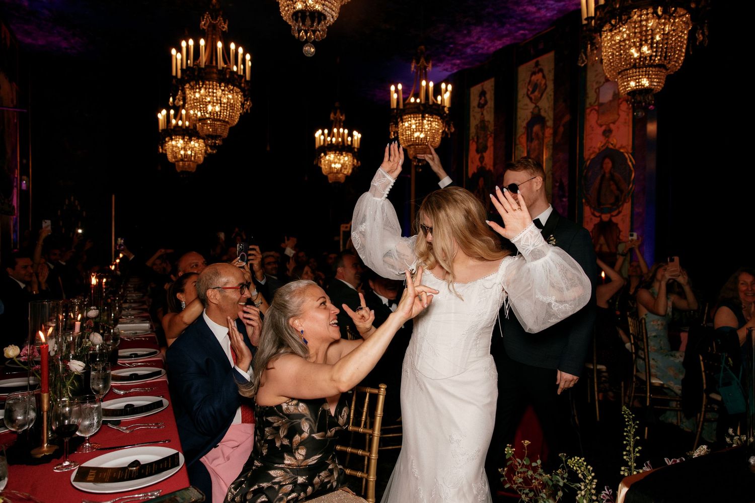 Bride and guest dancing joyfully at candlelit wedding reception under golden chandeliers.