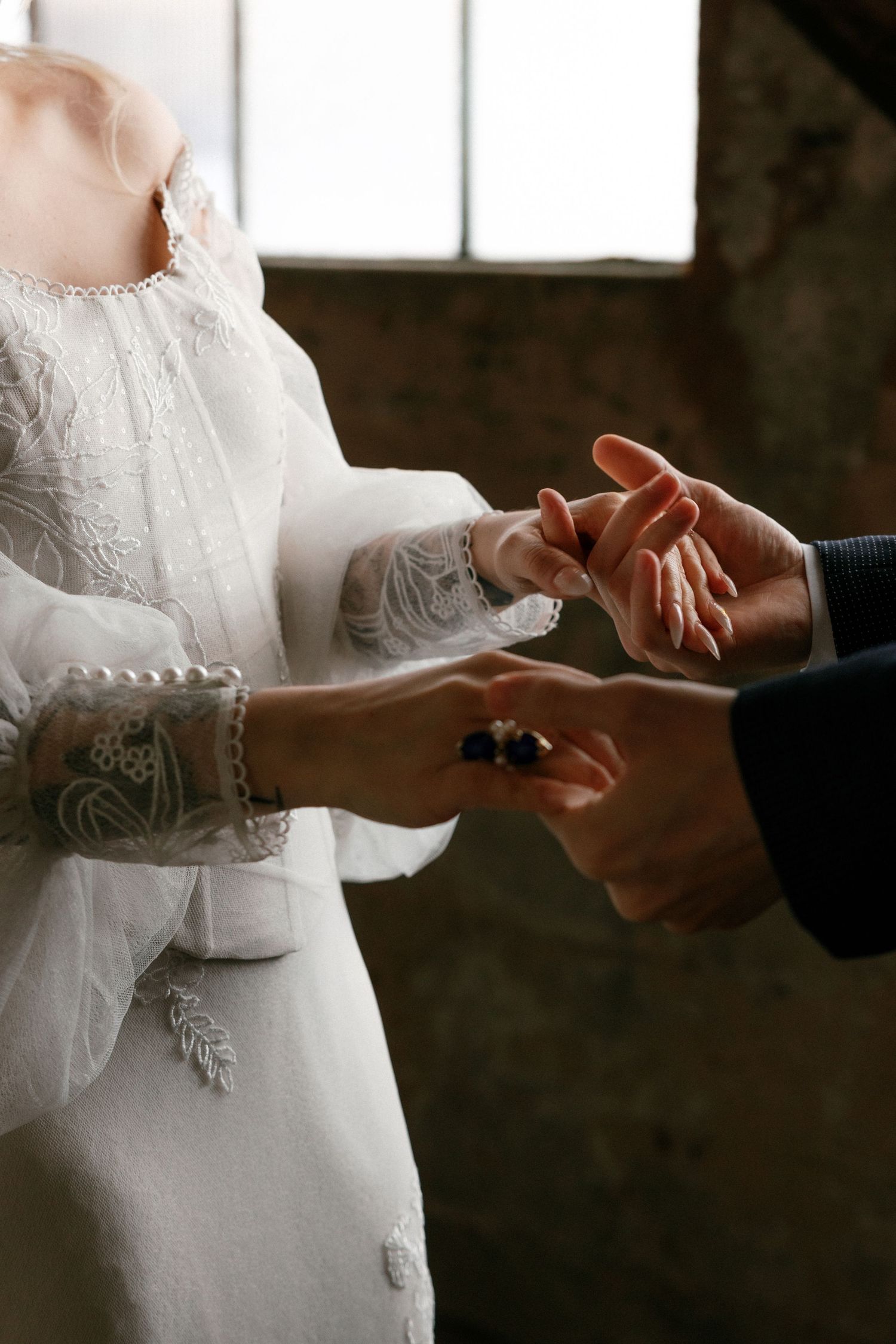 Close-up of bride and groom exchanging wedding rings, showcasing lace wedding dress sleeve and suit details.