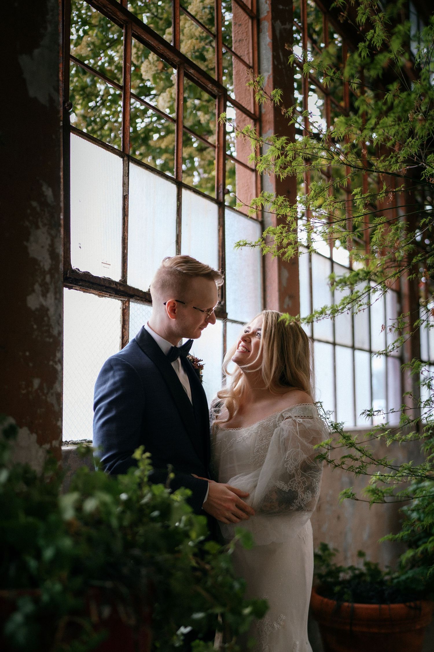 Bride and groom embrace tenderly surrounded by lush green vines near large industrial windows in a rustic wedding venue.