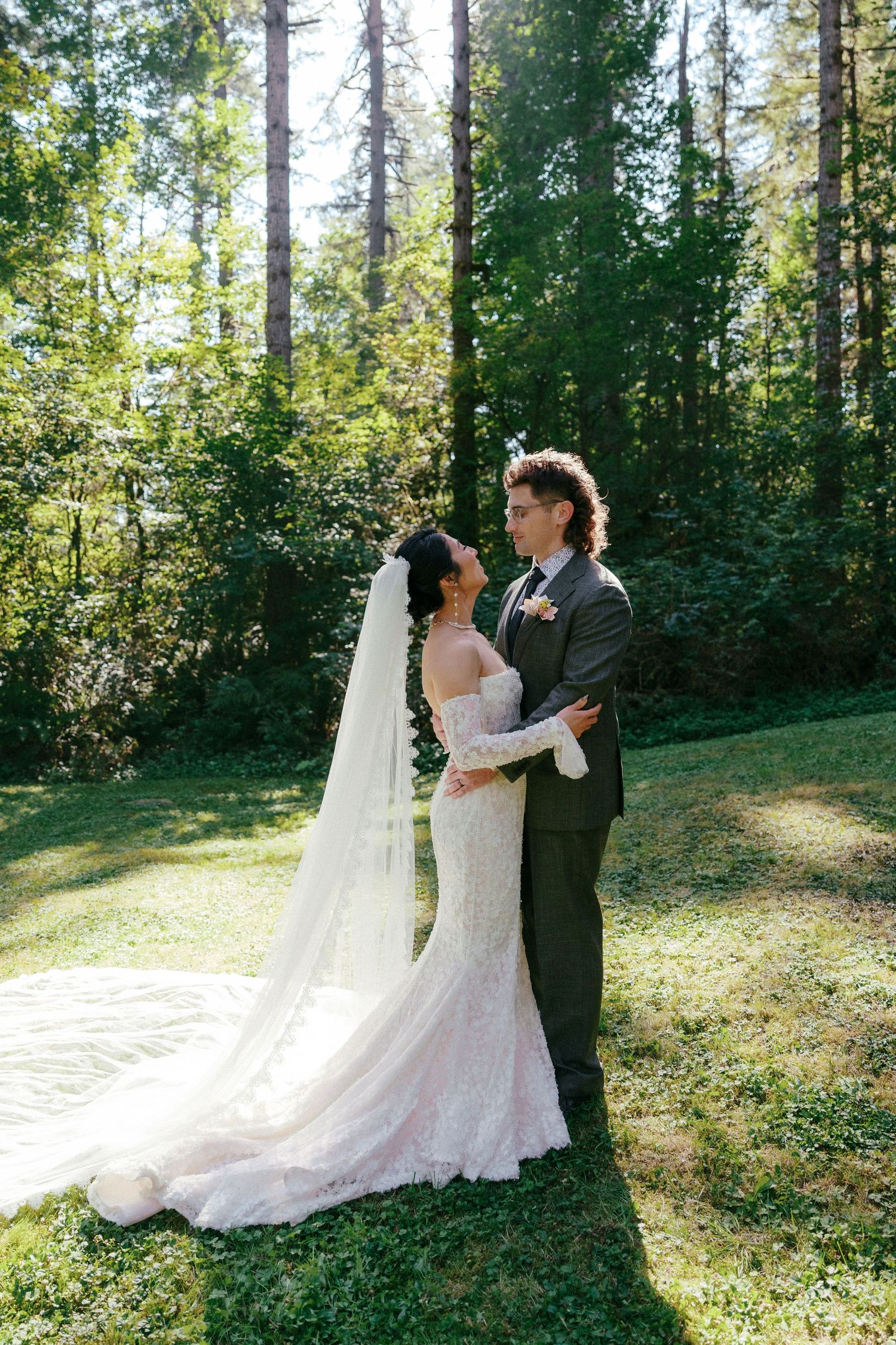 Bride and groom stand facing each other in full-length lace gown in forest.