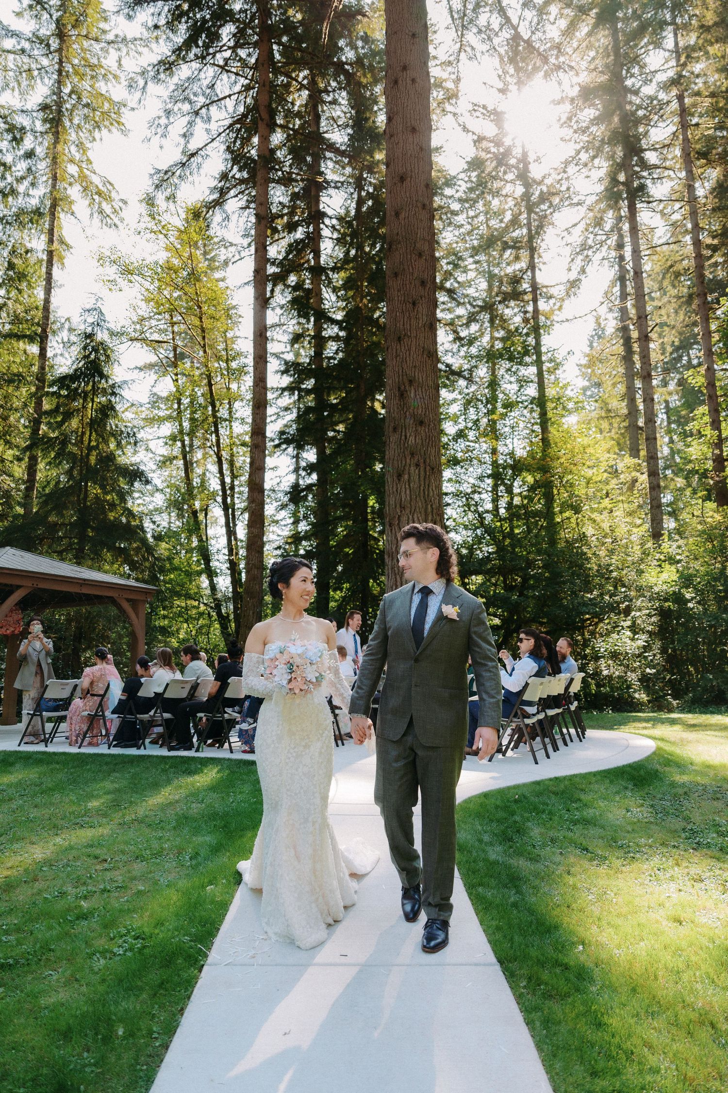 Bride and groom walk hand in hand down a sunlit outdoor aisle lined with tall evergreen trees.