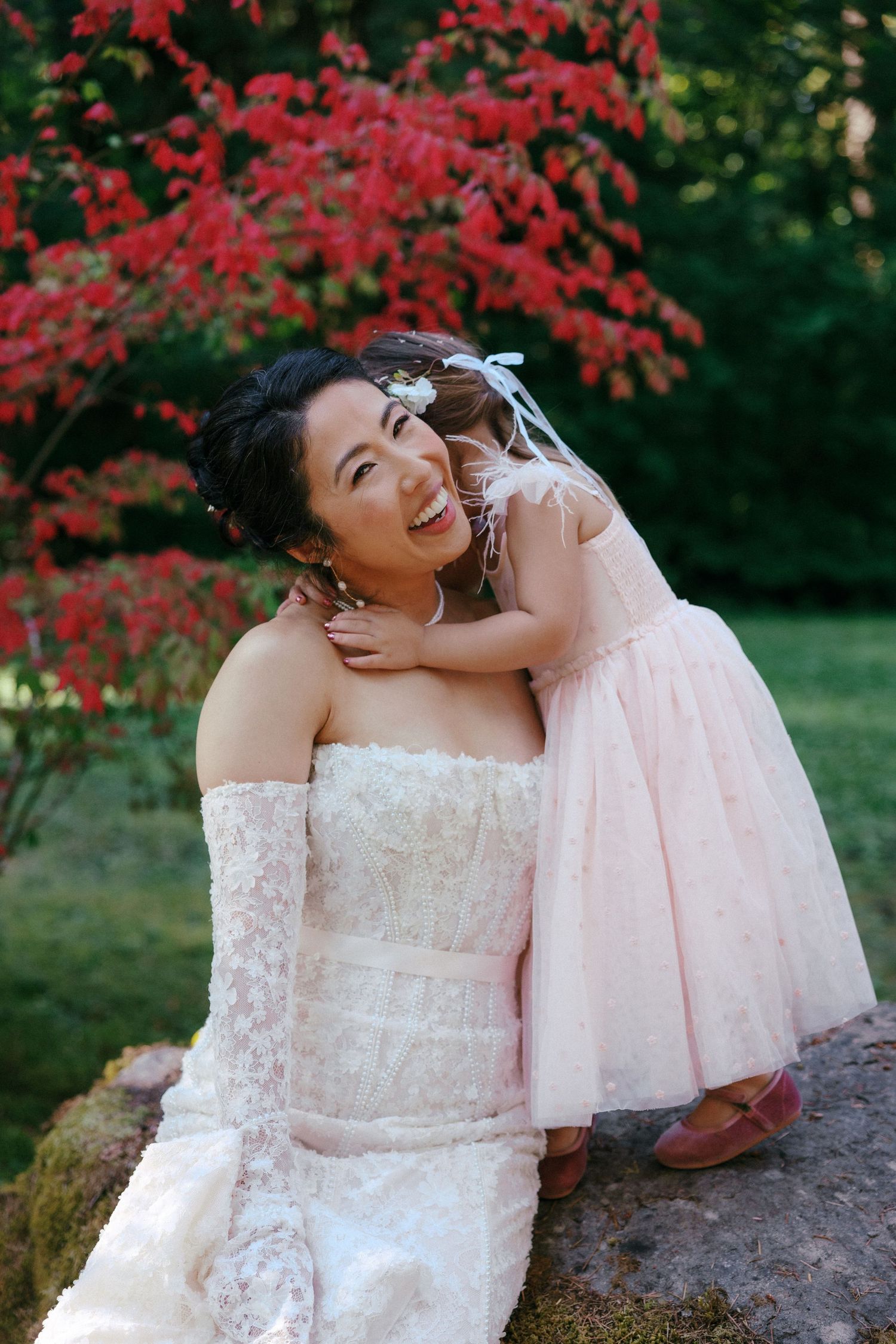 Smiling bride in strapless lace gown embraces a young flower girl in pink dress near red maple tree.