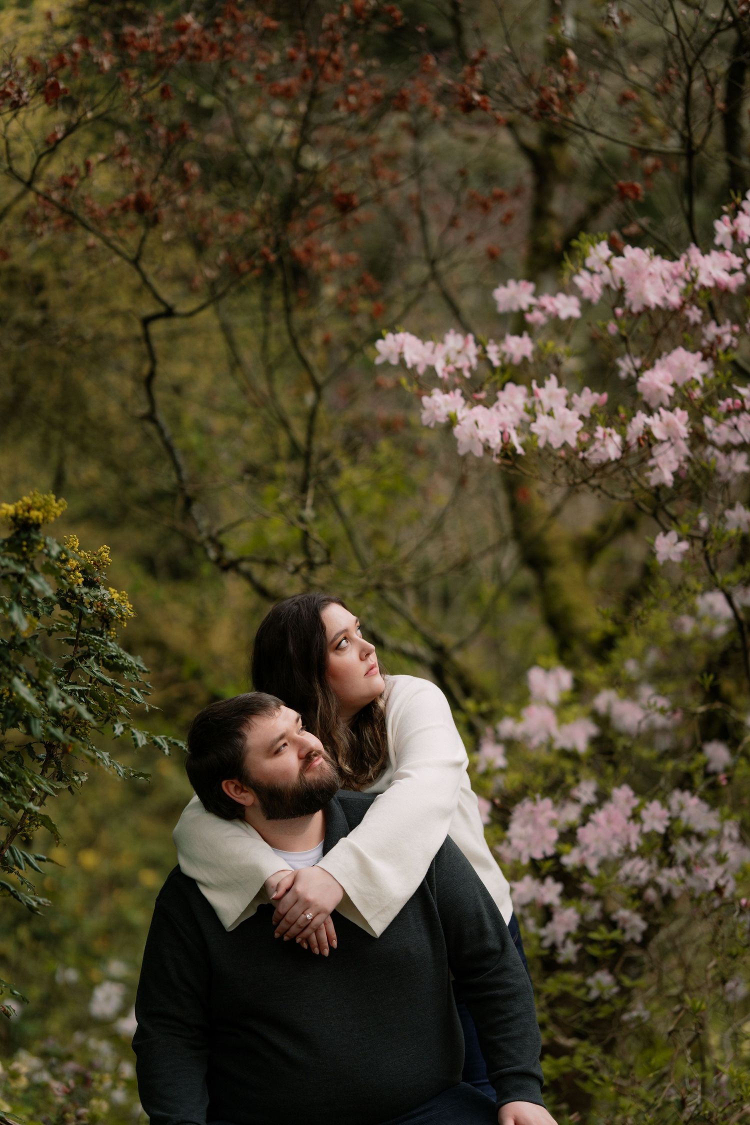 Woman hugging man from behind surrounded by pink rhododendrons in a forest setting.