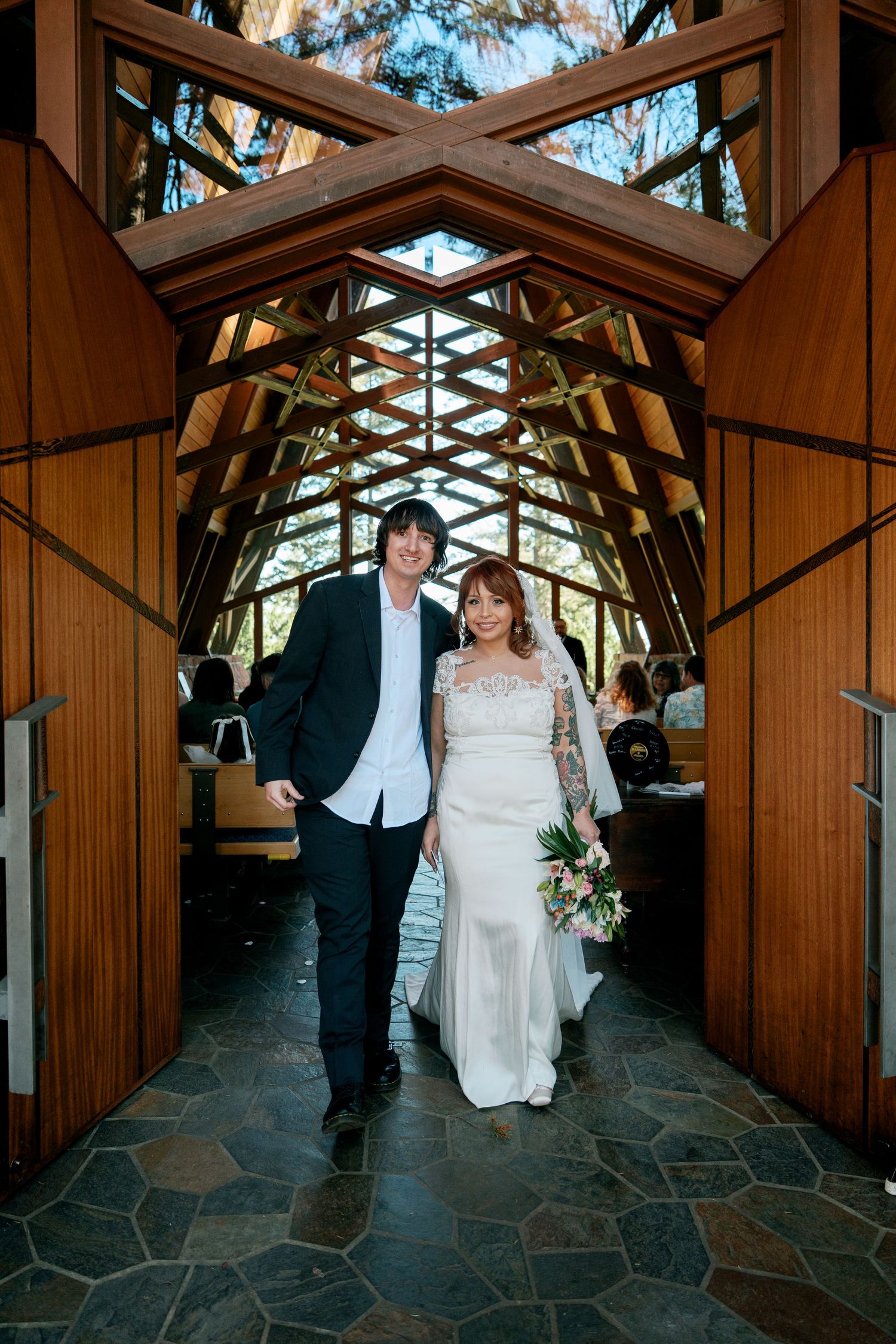 Bride and groom walk down the aisle of a stunning wooden chapel with geometric ceiling details.