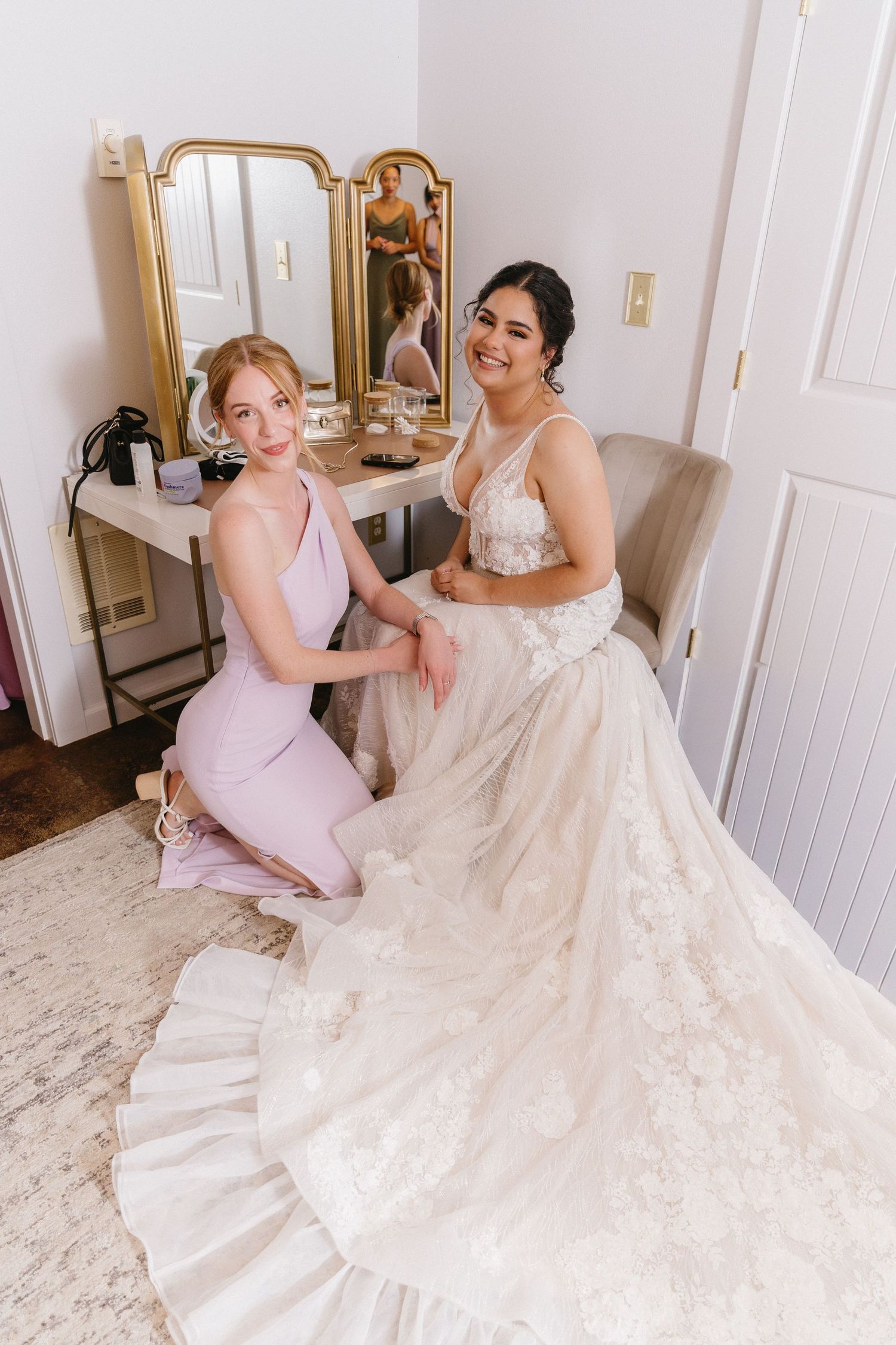 Bridesmaid in pink dress kneeling beside smiling bride in lace gown near vanity mirror.