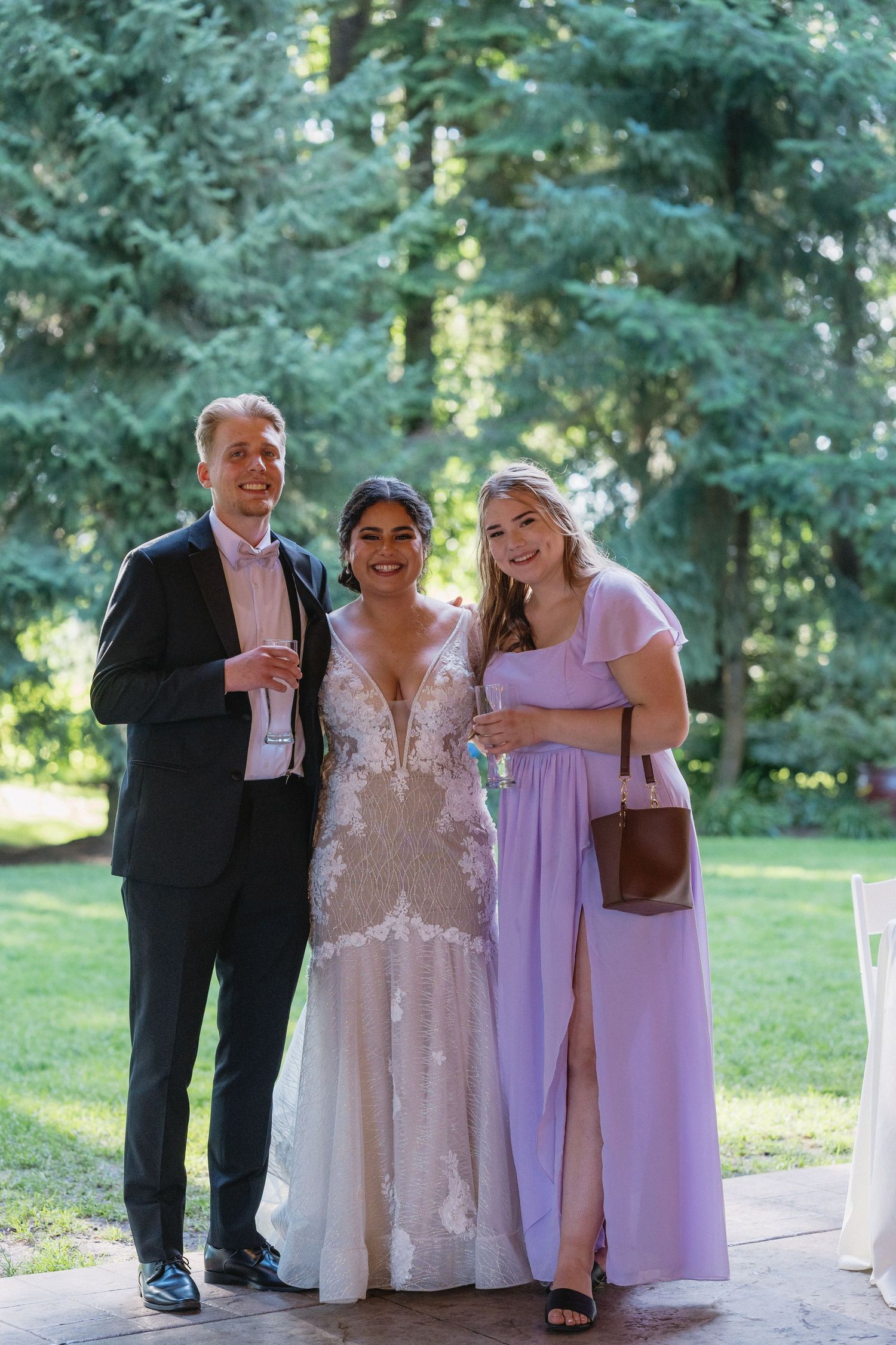 Bride in lace gown poses with two smiling guests in a sunlit outdoor wedding venue.