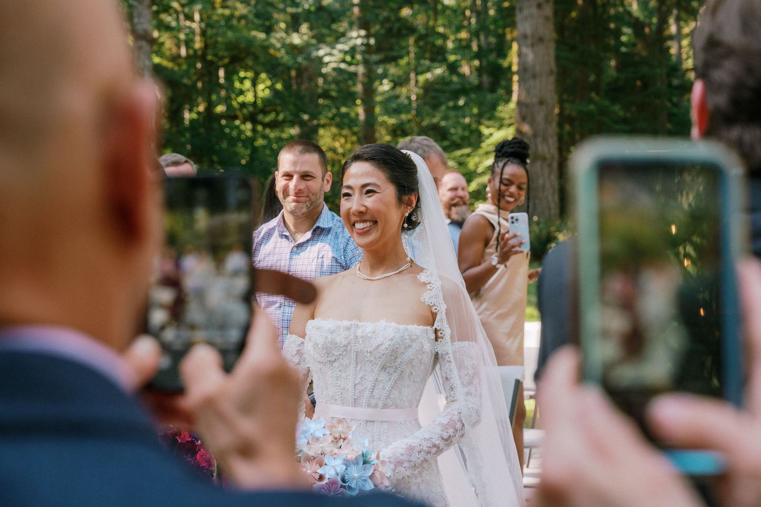 Joyful Asian bride in lace gown laughs as guests photograph her during outdoor forest wedding.