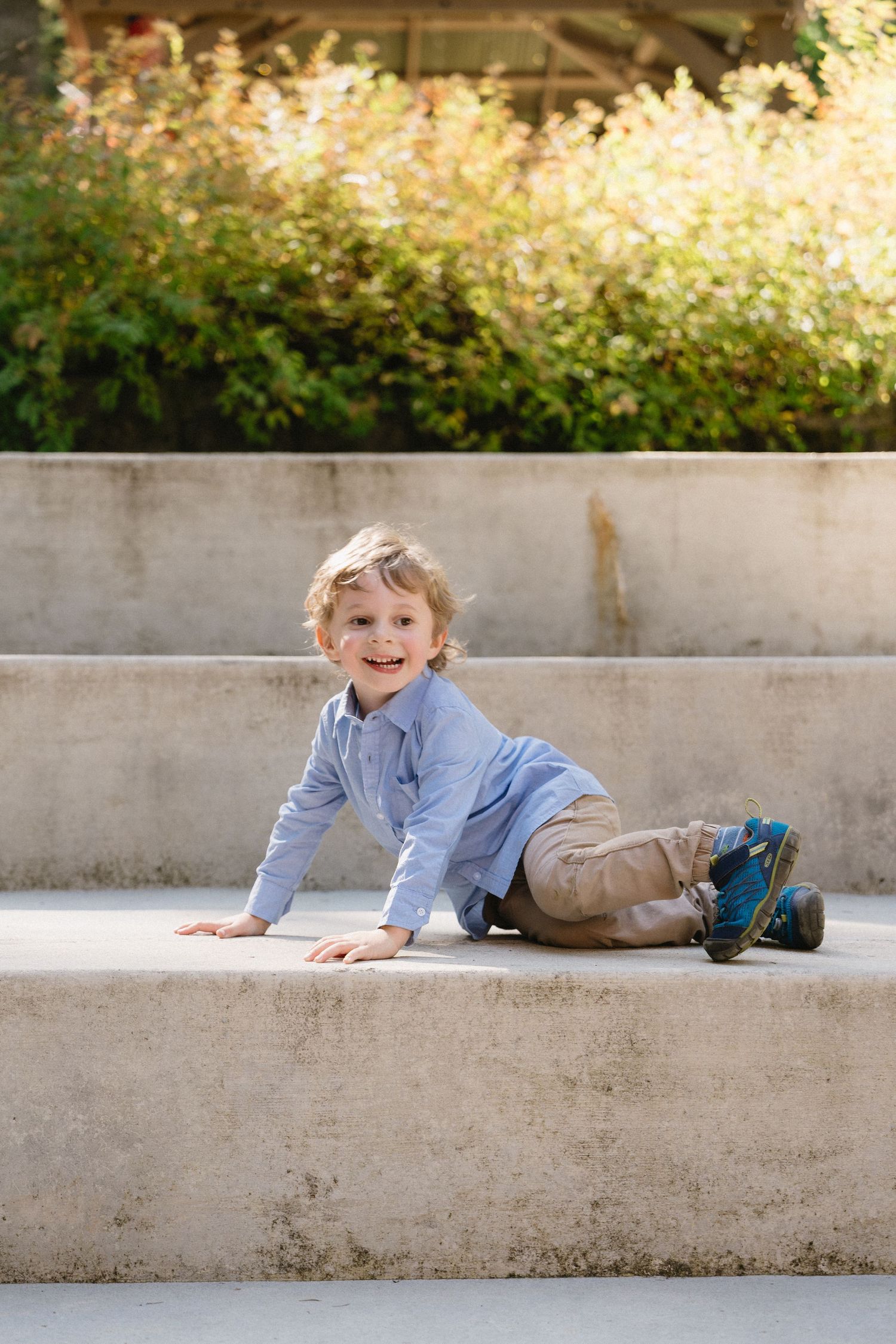 A smiling young blond boy in a blue shirt and khaki pants sits on concrete outdoor steps.