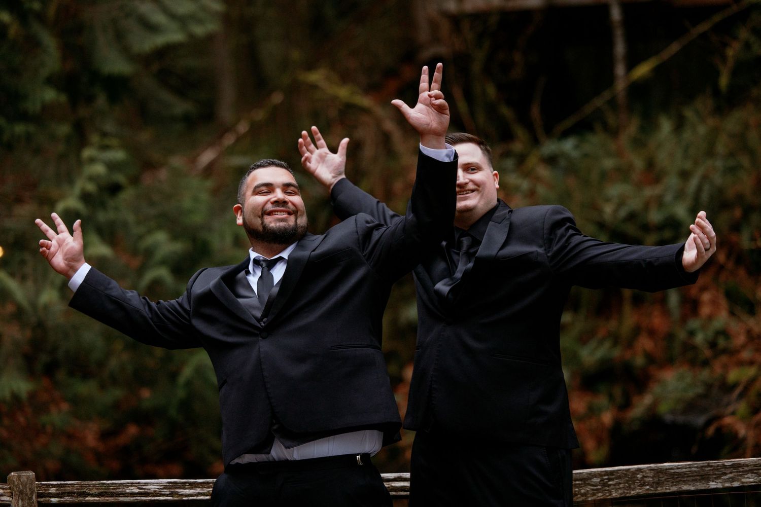 Two grooms in black suits celebrating with arms wide open in a forest wedding photo.