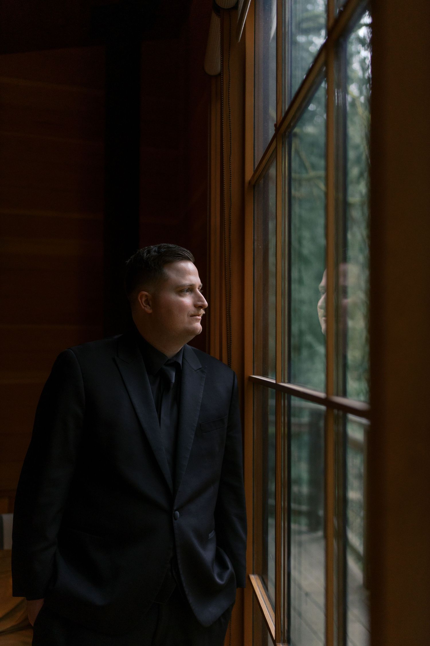 Groom in black suit gazes thoughtfully out large wooden-framed windows in dimly lit interior.
