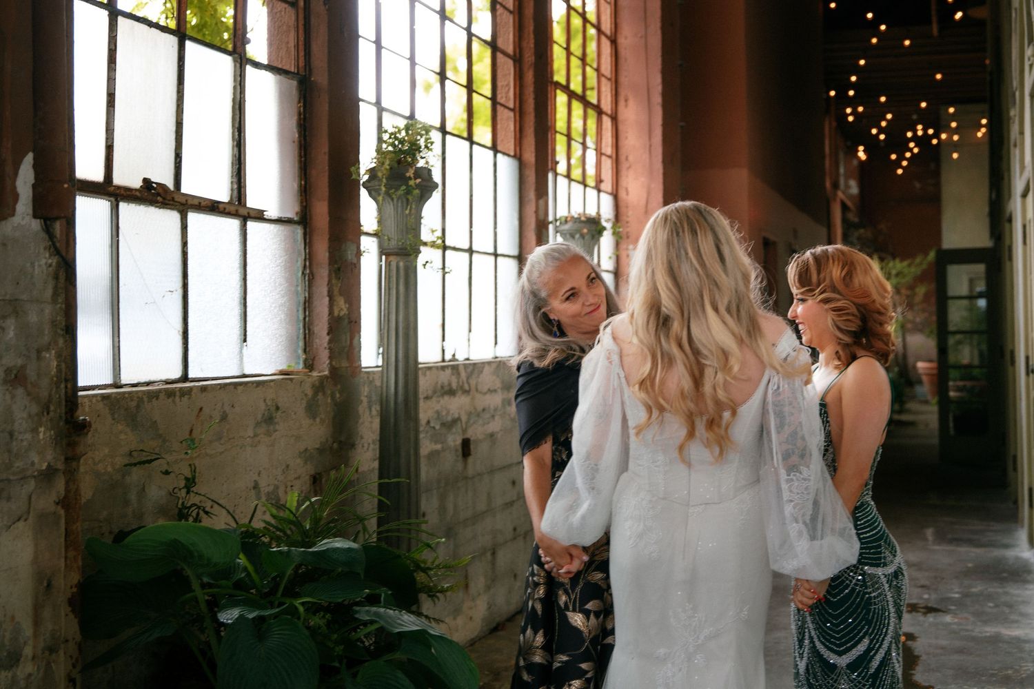 Bride in white lace gown chatting with guests near large windows in a rustic industrial wedding venue.