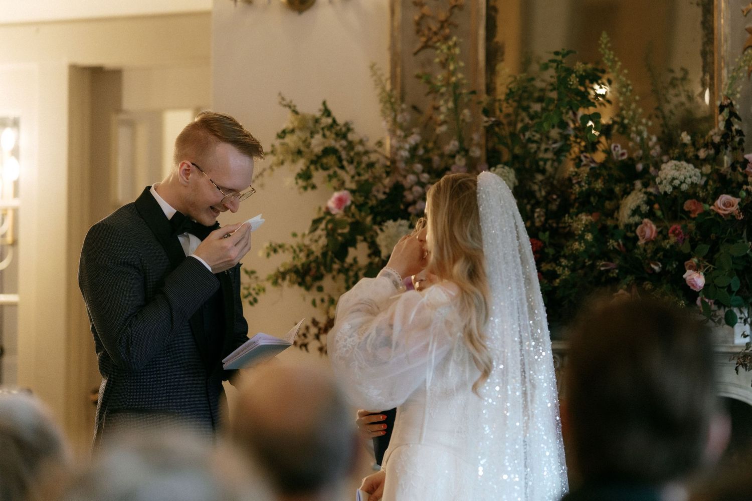 Emotional bride and groom exchange vows before lush floral arrangement at indoor wedding ceremony.