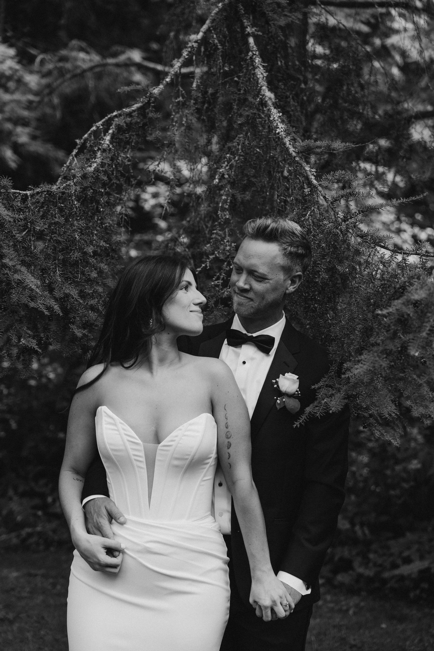 Black and white photo of bride in strapless gown and groom in tuxedo standing before evergreen trees.