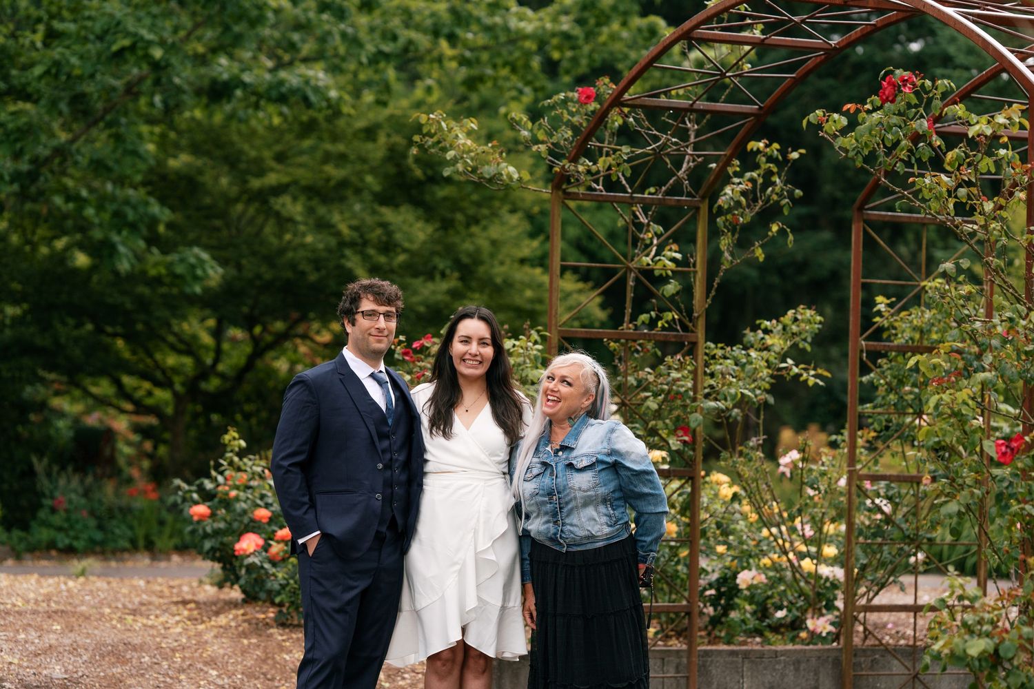Couple in wedding attire poses with older woman in rose garden with metal arbor backdrop.