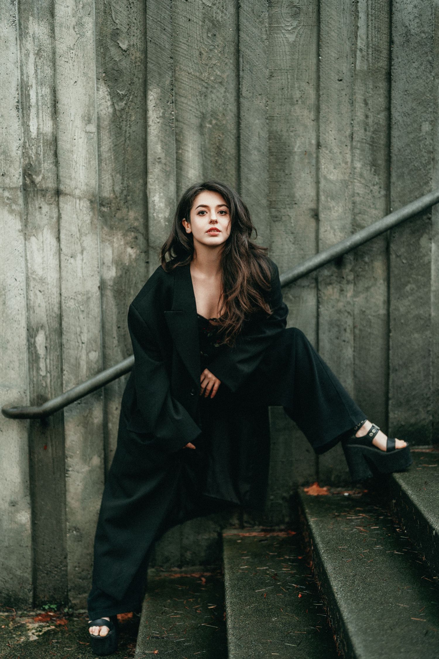 Young woman in black coat and wide-leg pants poses on concrete steps against gray wall.