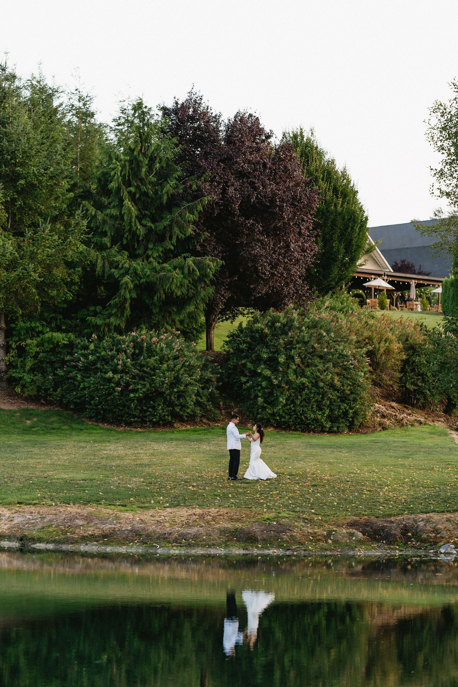 Wedding couple holds hands by a serene pond with lush greenery and a venue building reflected in water.