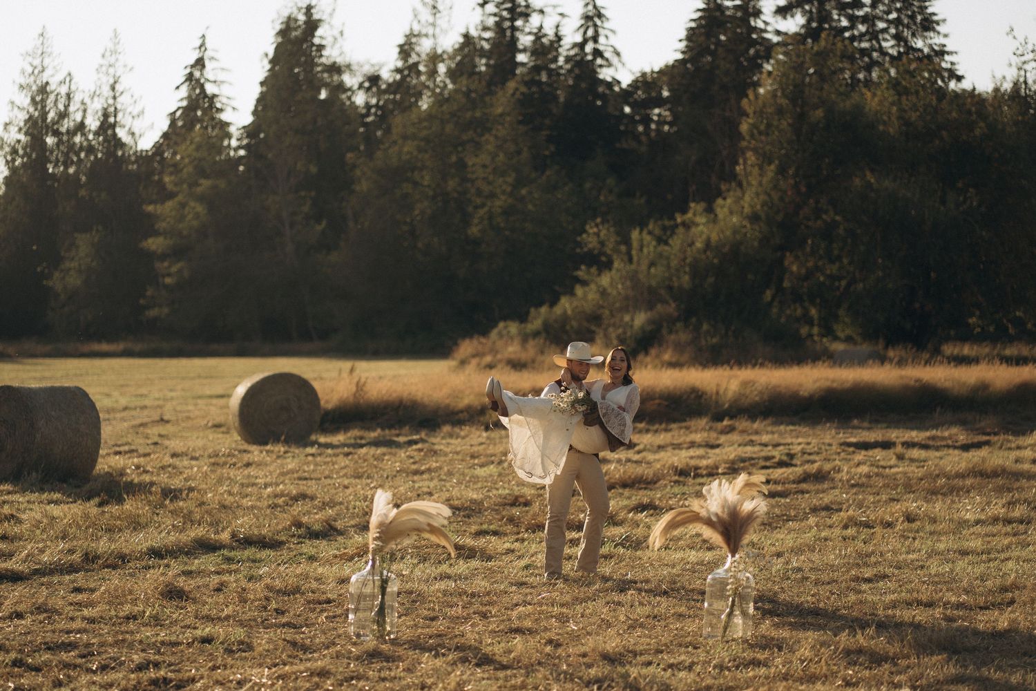 Groom lifts bride joyfully in a sunlit field with hay bales and pampas grass at a rustic outdoor wedding.