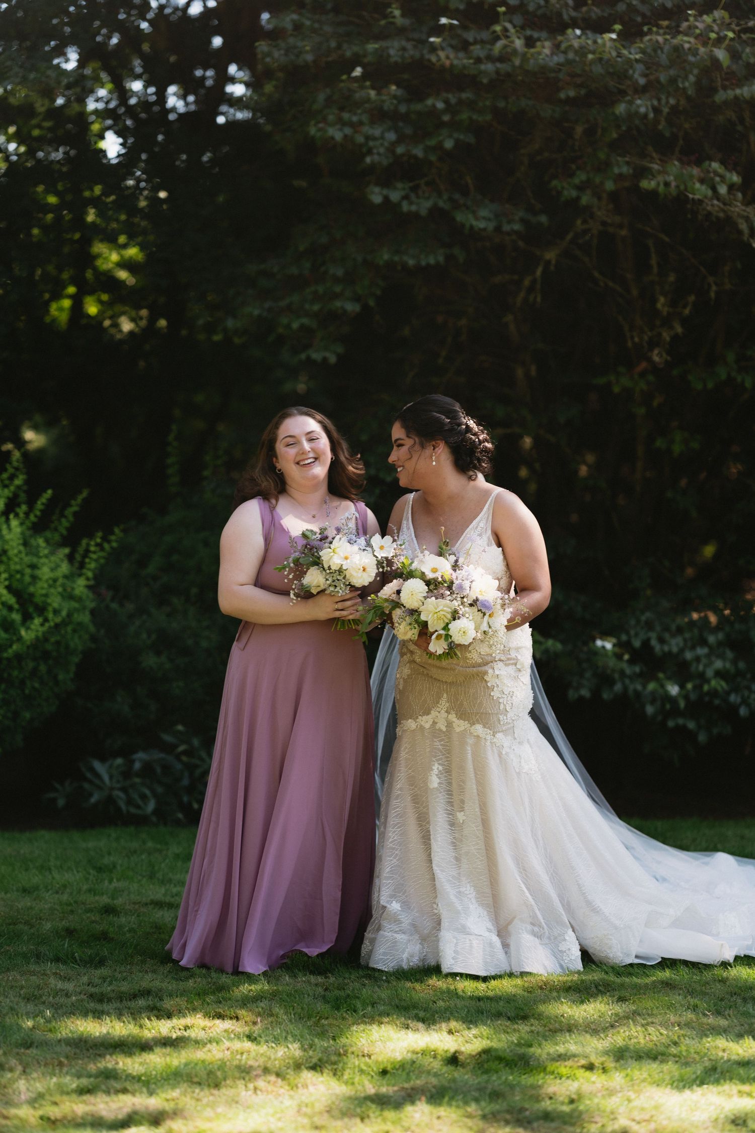 Bride and bridesmaid in mauve dress laughing together outdoors holding white floral bouquets.
