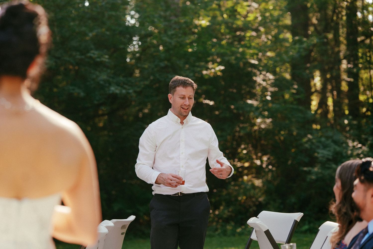 Man in white shirt speaks at an outdoor wedding ceremony surrounded by lush green trees at golden hour.