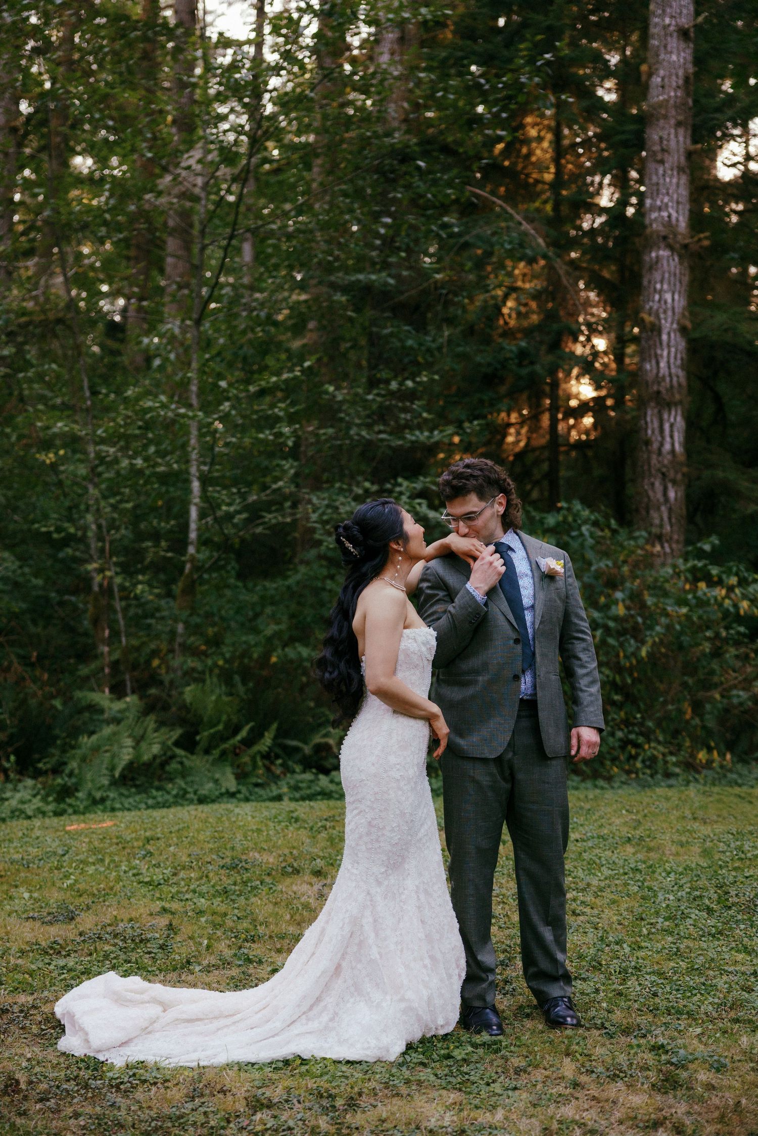 Groom in gray suit kisses bride's hand at sunset in a forested outdoor wedding portrait.