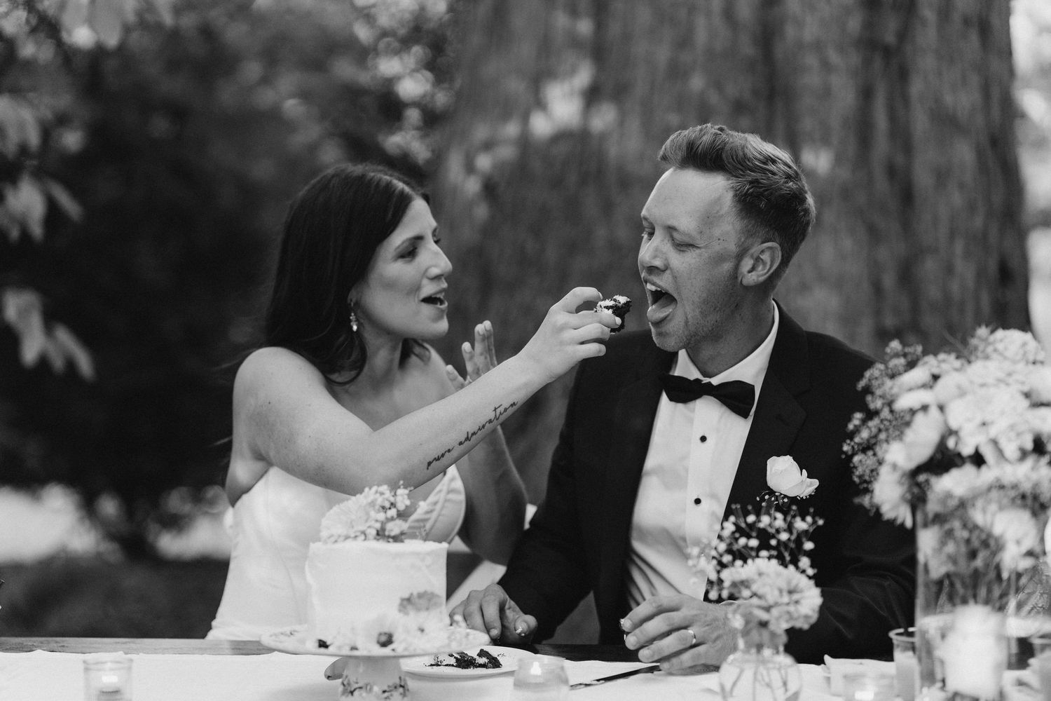 Black and white photo of bride feeding wedding cake to groom at reception table.