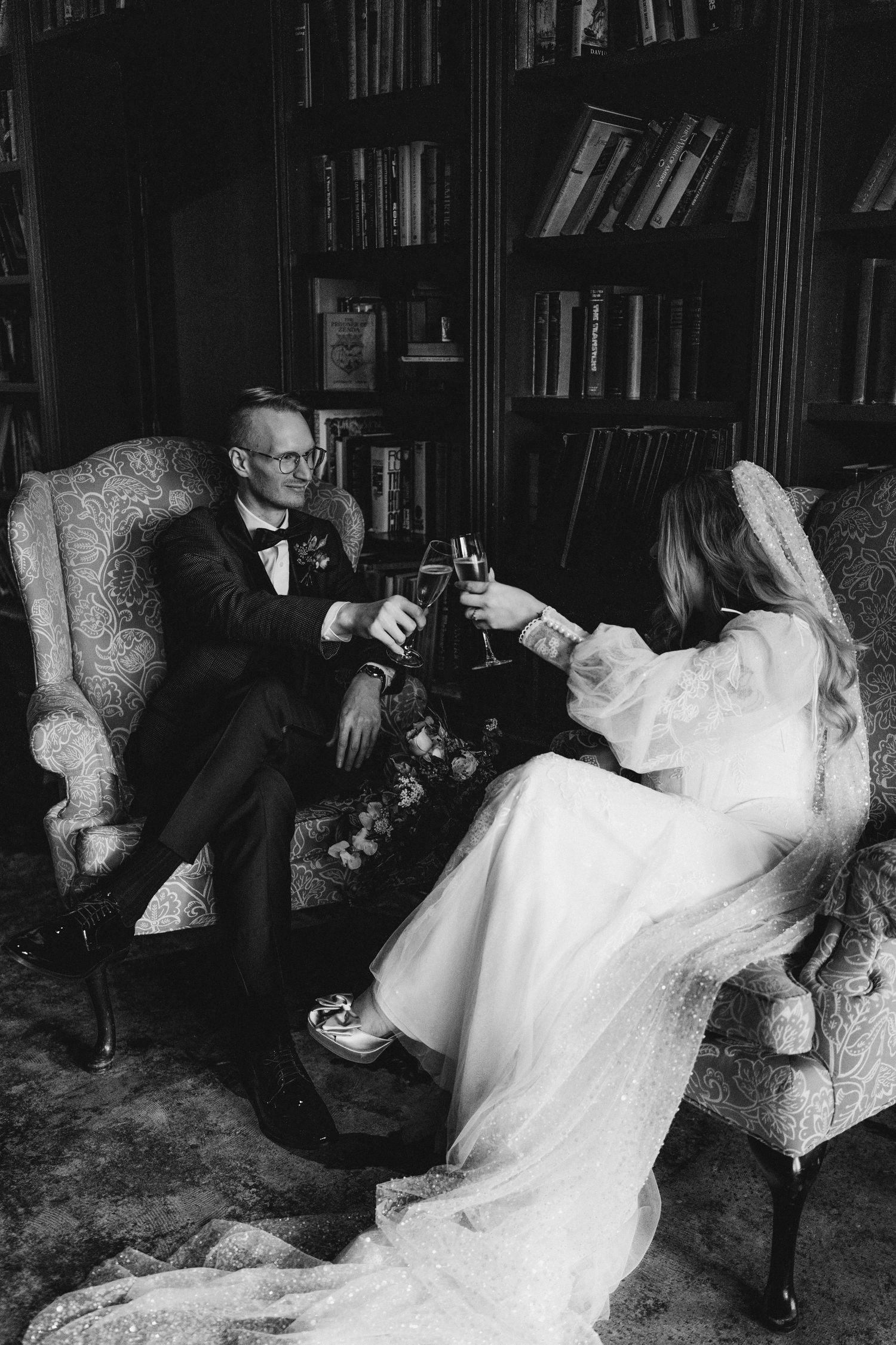Bride and groom toasting champagne in a dark library with bookshelves, black and white wedding photo.