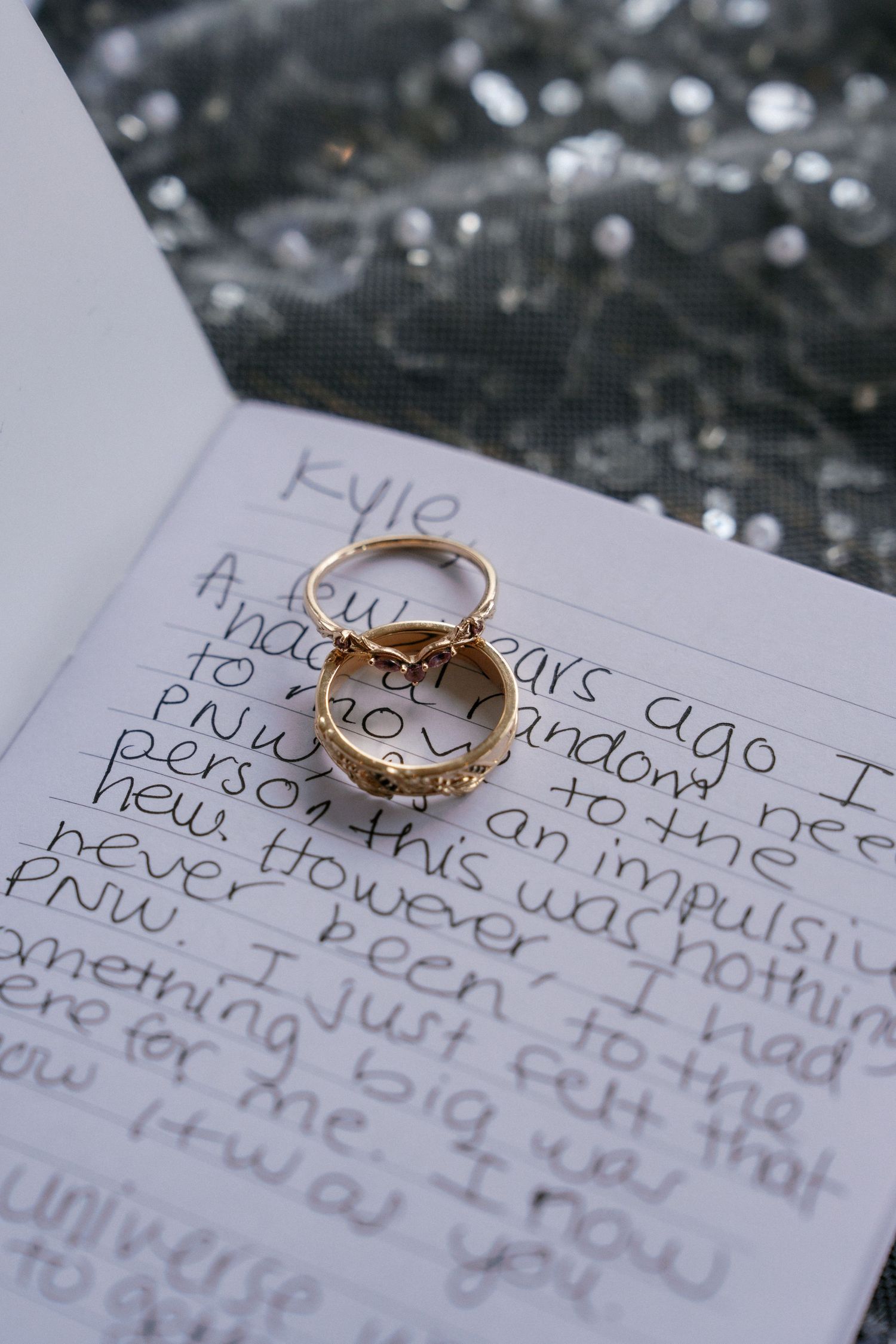 Gold wedding rings resting on handwritten vows notebook with sequined fabric background.