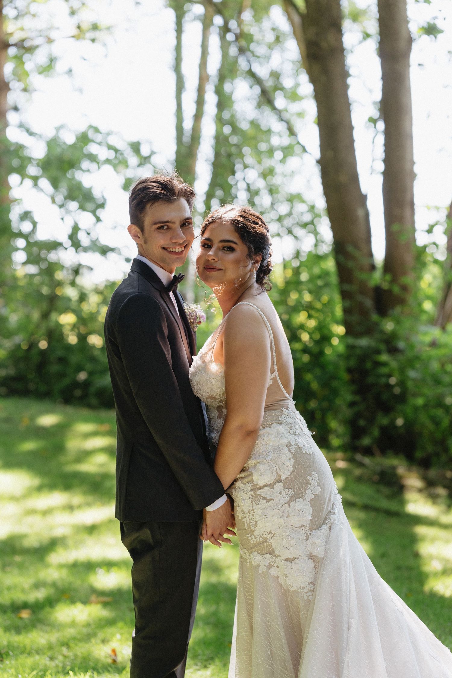 Smiling bride and groom pose together in a sunny garden, bride wearing floral lace wedding gown.