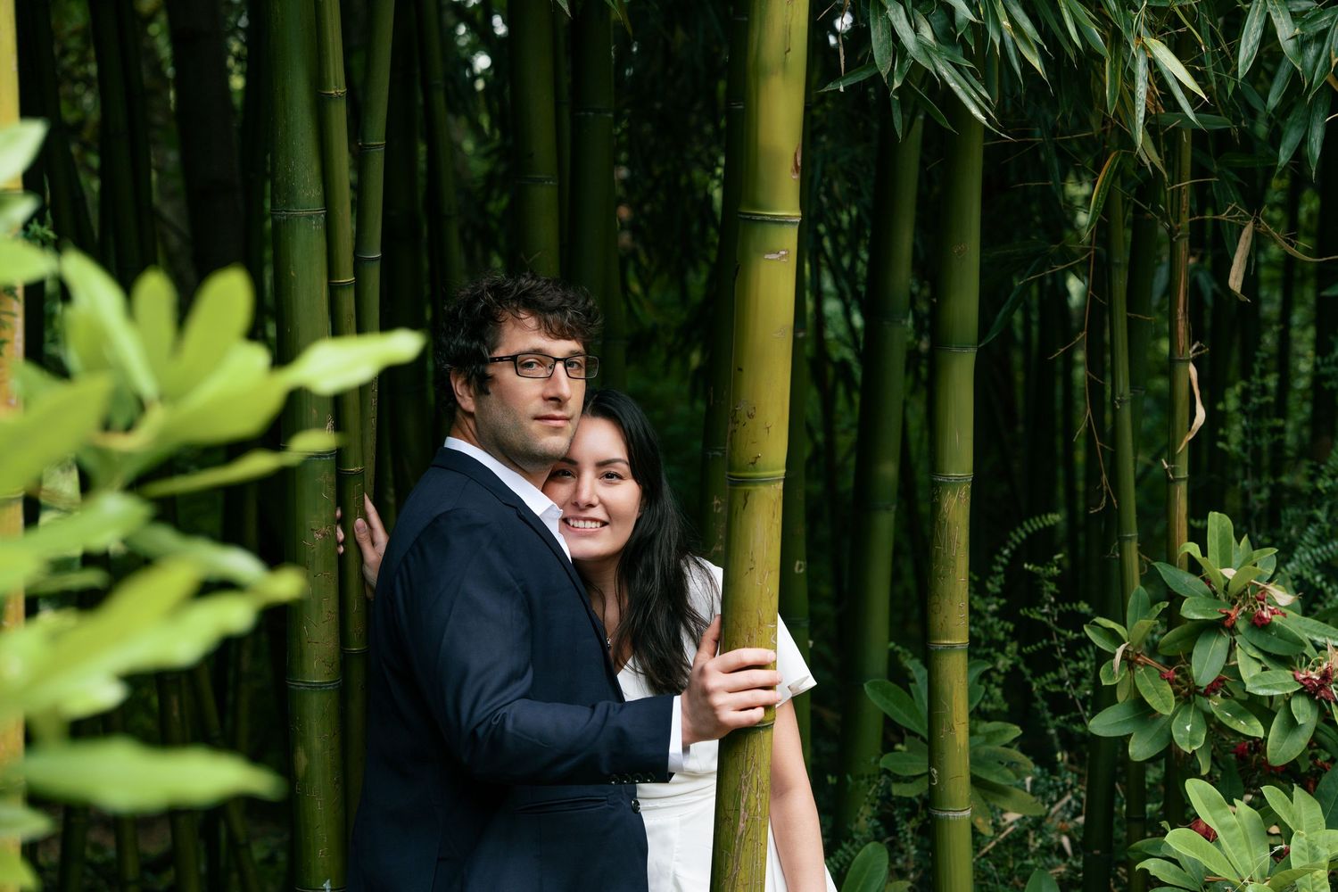 Couple embracing and smiling among tall bamboo stalks in a lush garden, woman in white dress and man in suit.