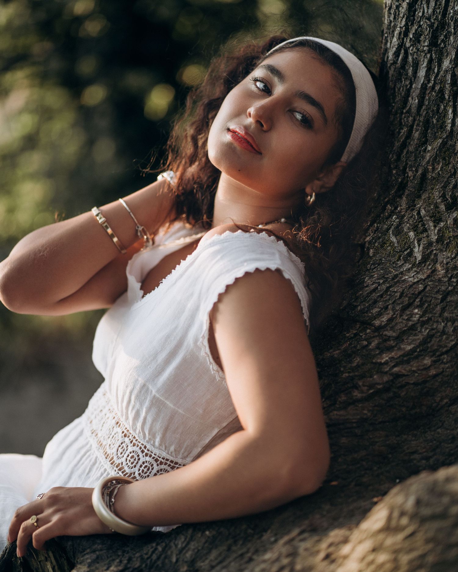 Young woman in white lace dress leaning against tree trunk, wearing gold jewelry and white headband.