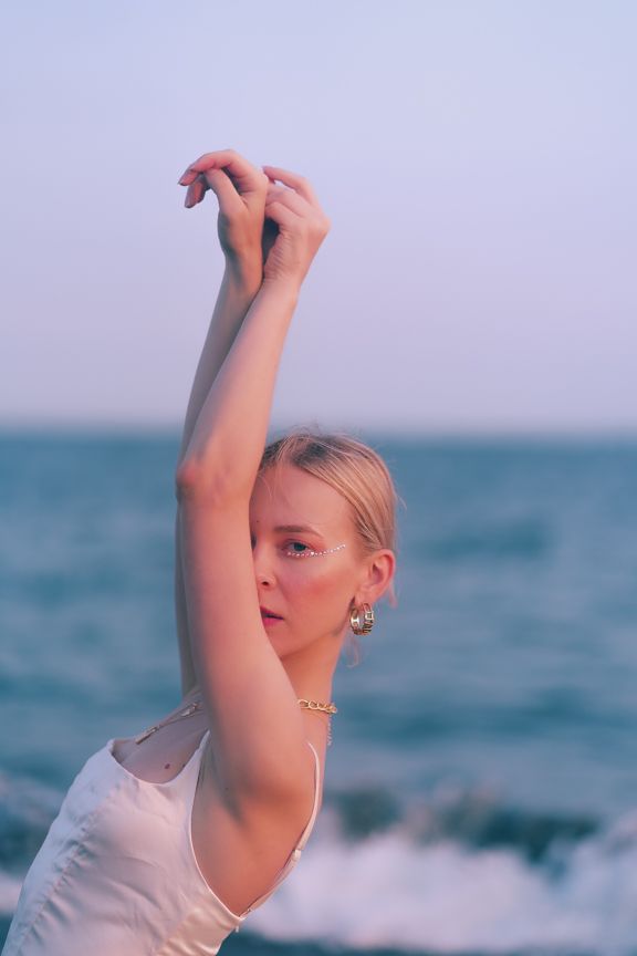 Blonde woman in white tank top poses with arms raised at beach during sunset.