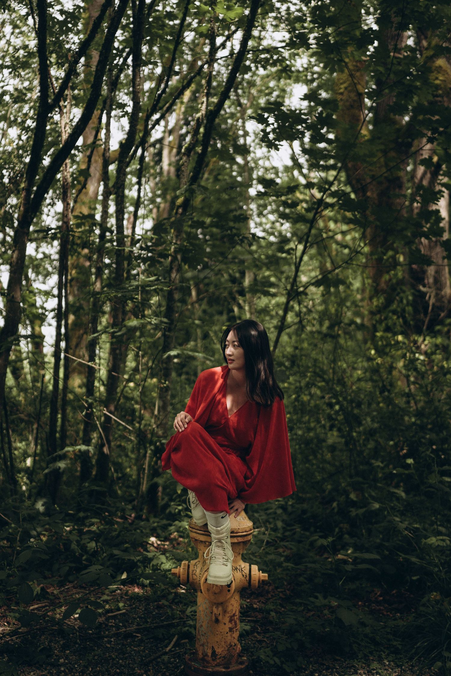 Woman in red dress perched atop a yellow fire hydrant in a lush green forest.