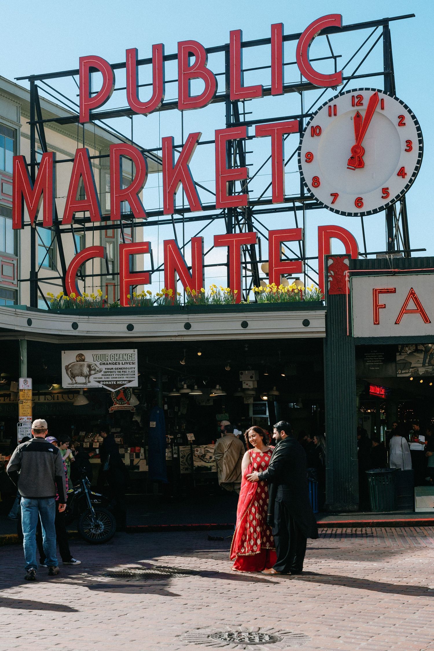 South Asian couple in red and black traditional wear posing under Seattle's Public Market Center sign.