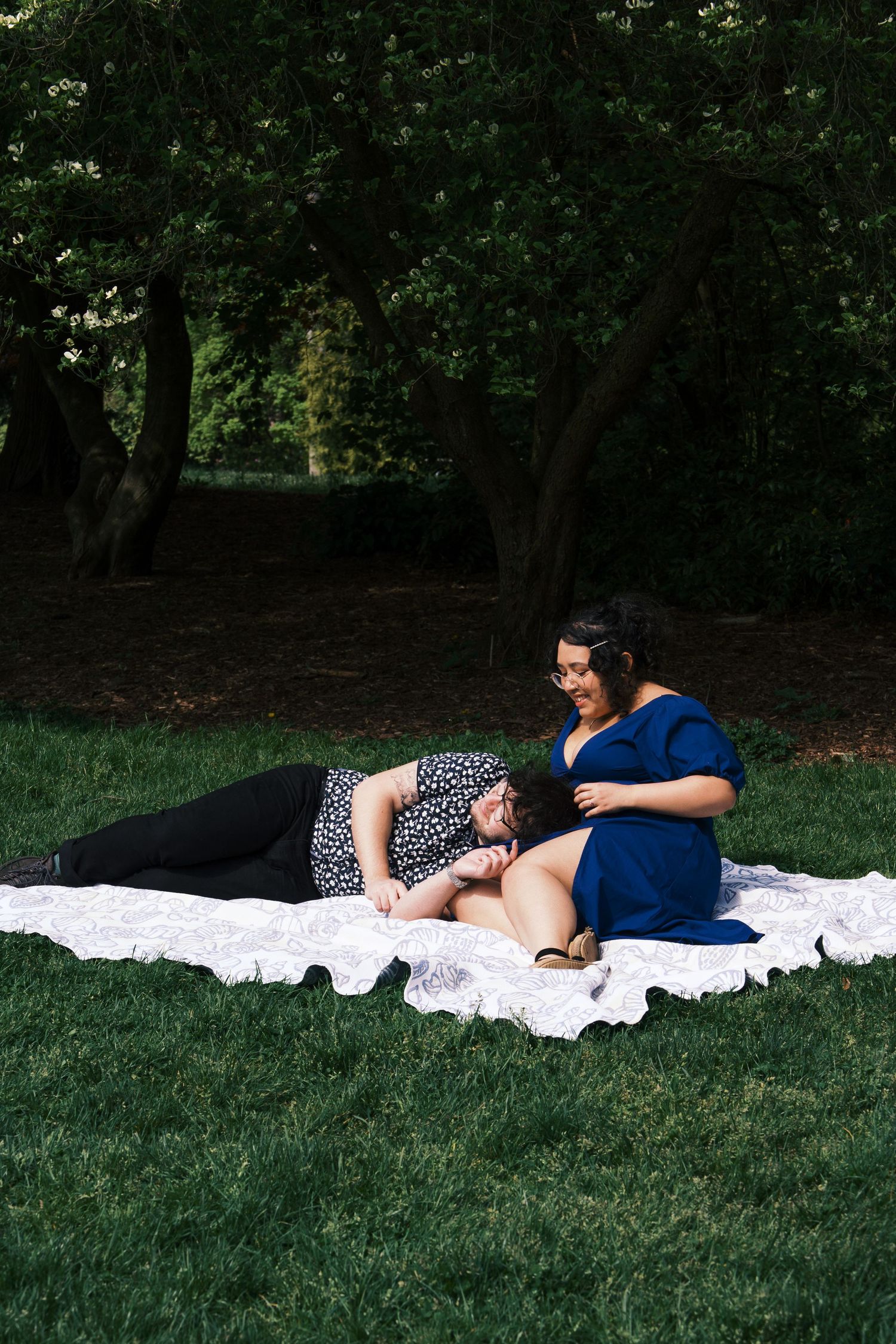 Couple relaxing on a white blanket on green grass, one laying down, the other sitting in a blue dress.