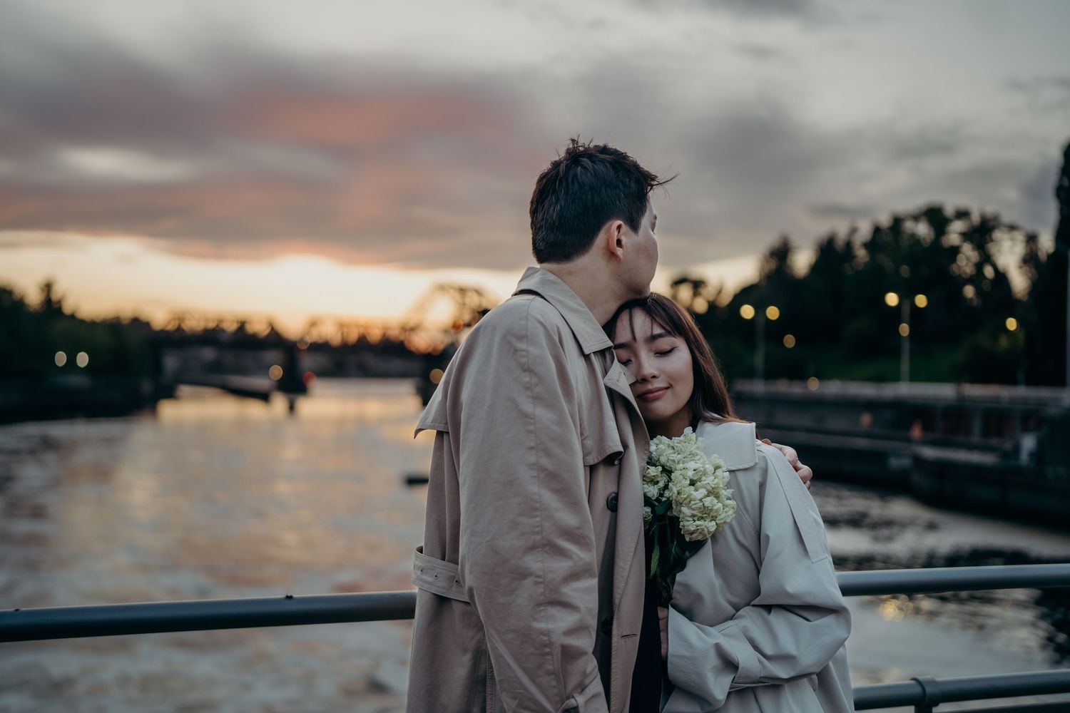 Romantic couple embracing at sunset on a bridge, woman holding white flowers by a river.