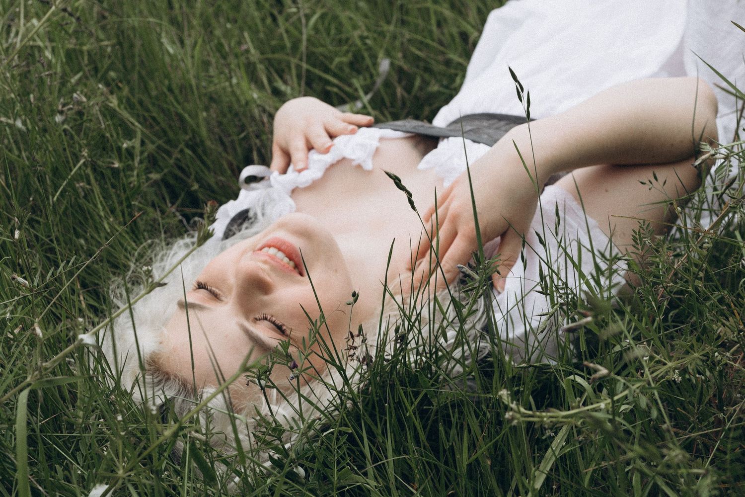 Young woman in white dress lying peacefully in tall green grass, smiling softly.